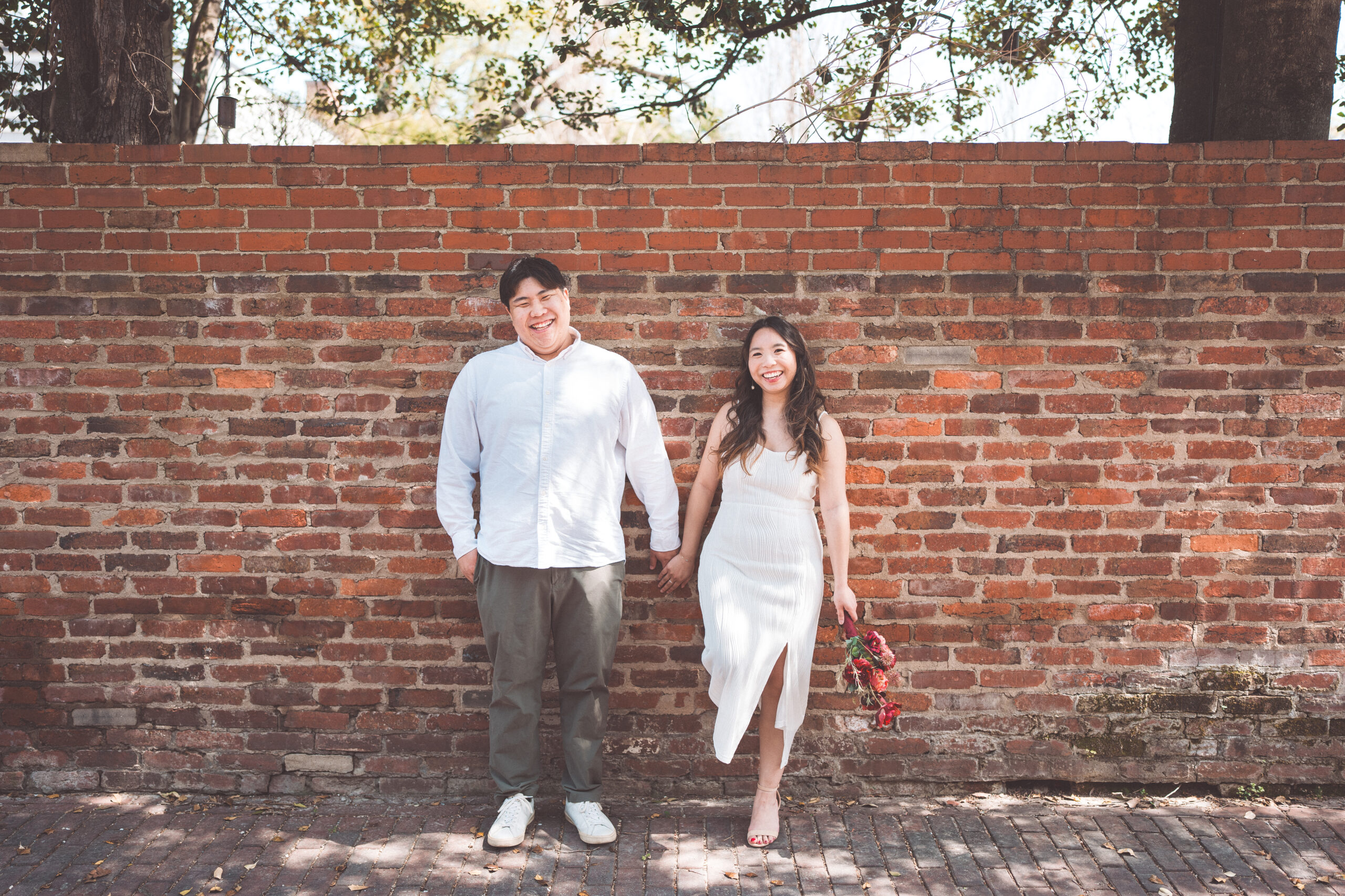 Playful couple holding hands and laughing in front of a historic brick wall during an engagement session