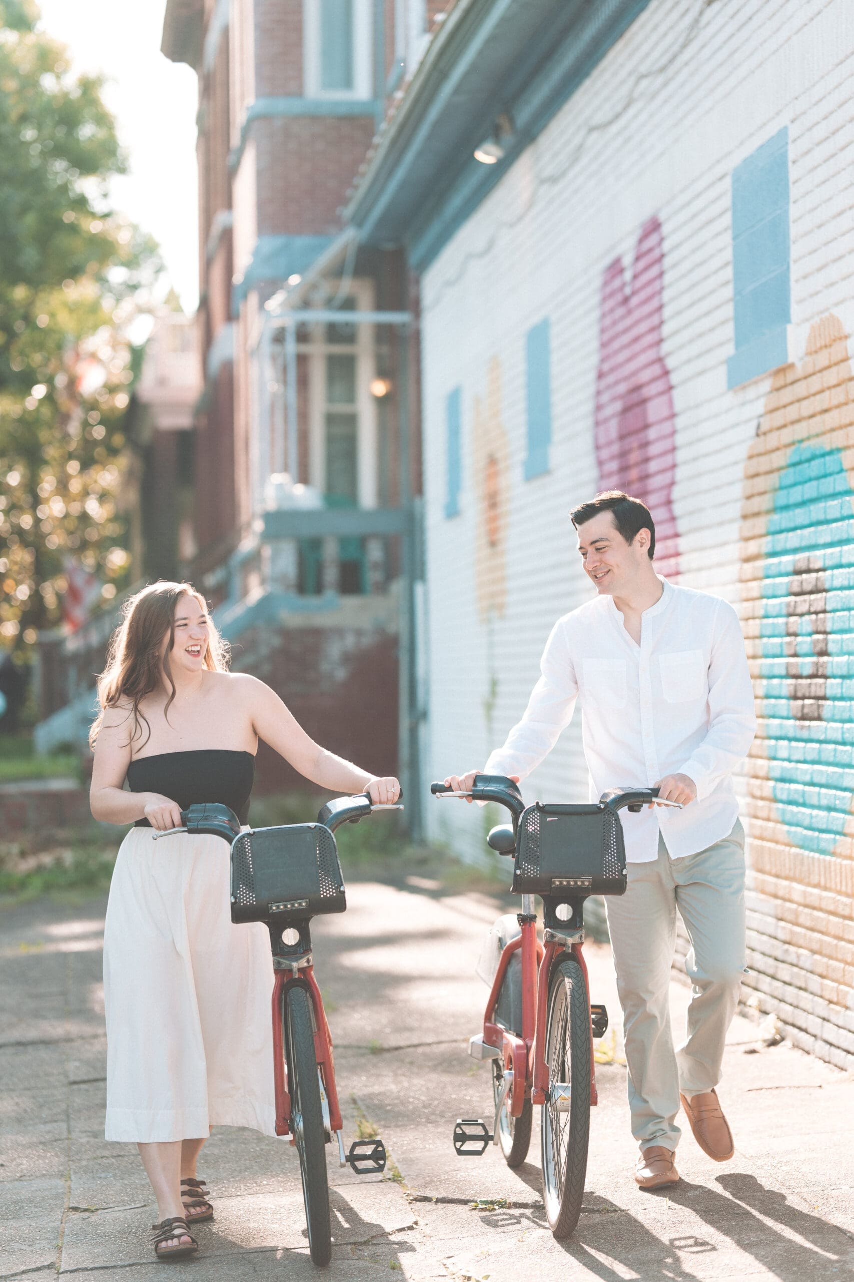 A couple walking their bikes alongside each other on the sidewalk with a colorful mural behind them.