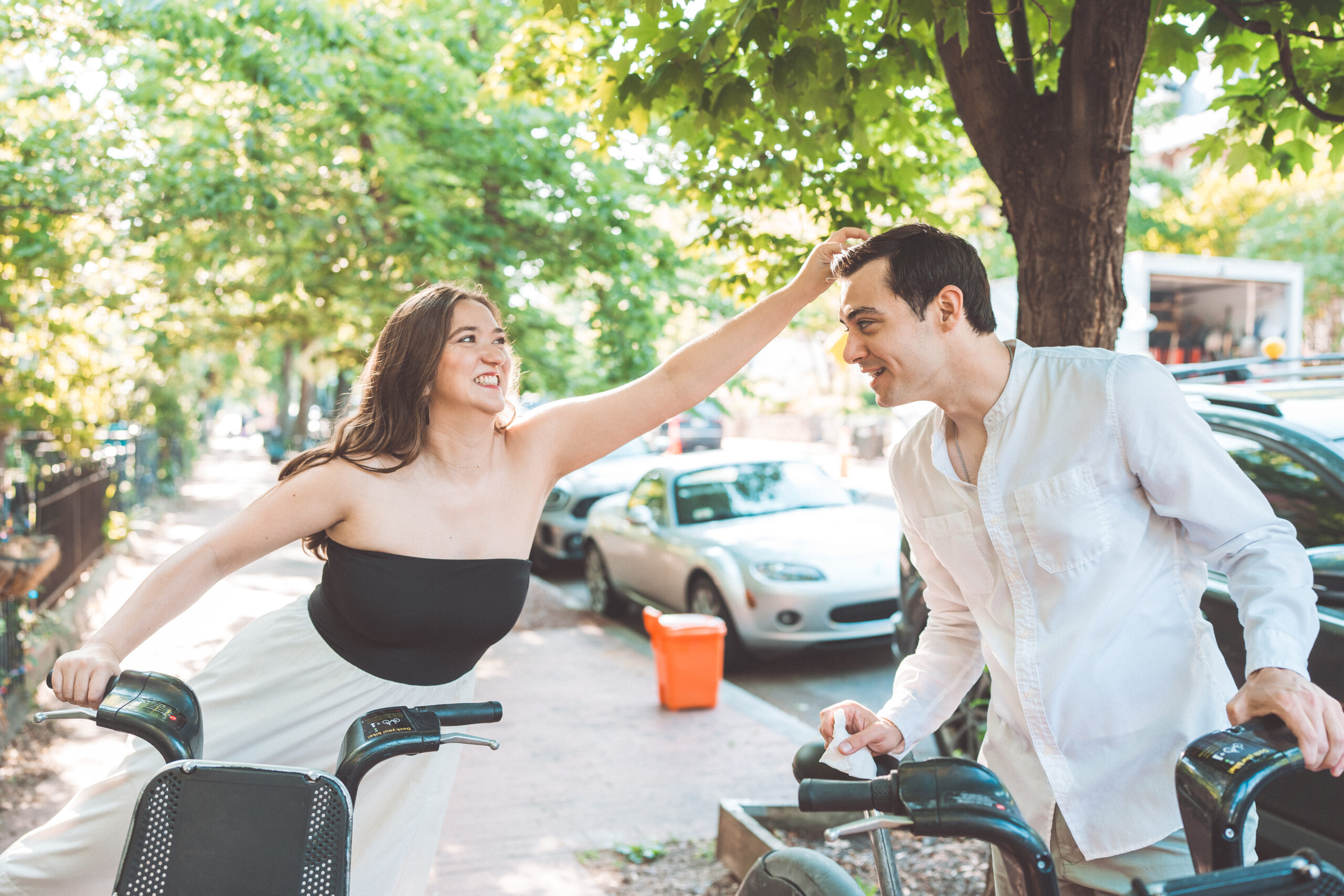 Playful candid of a couple laughing together with their bikes on a tree-lined Washington DC street during engagement photos