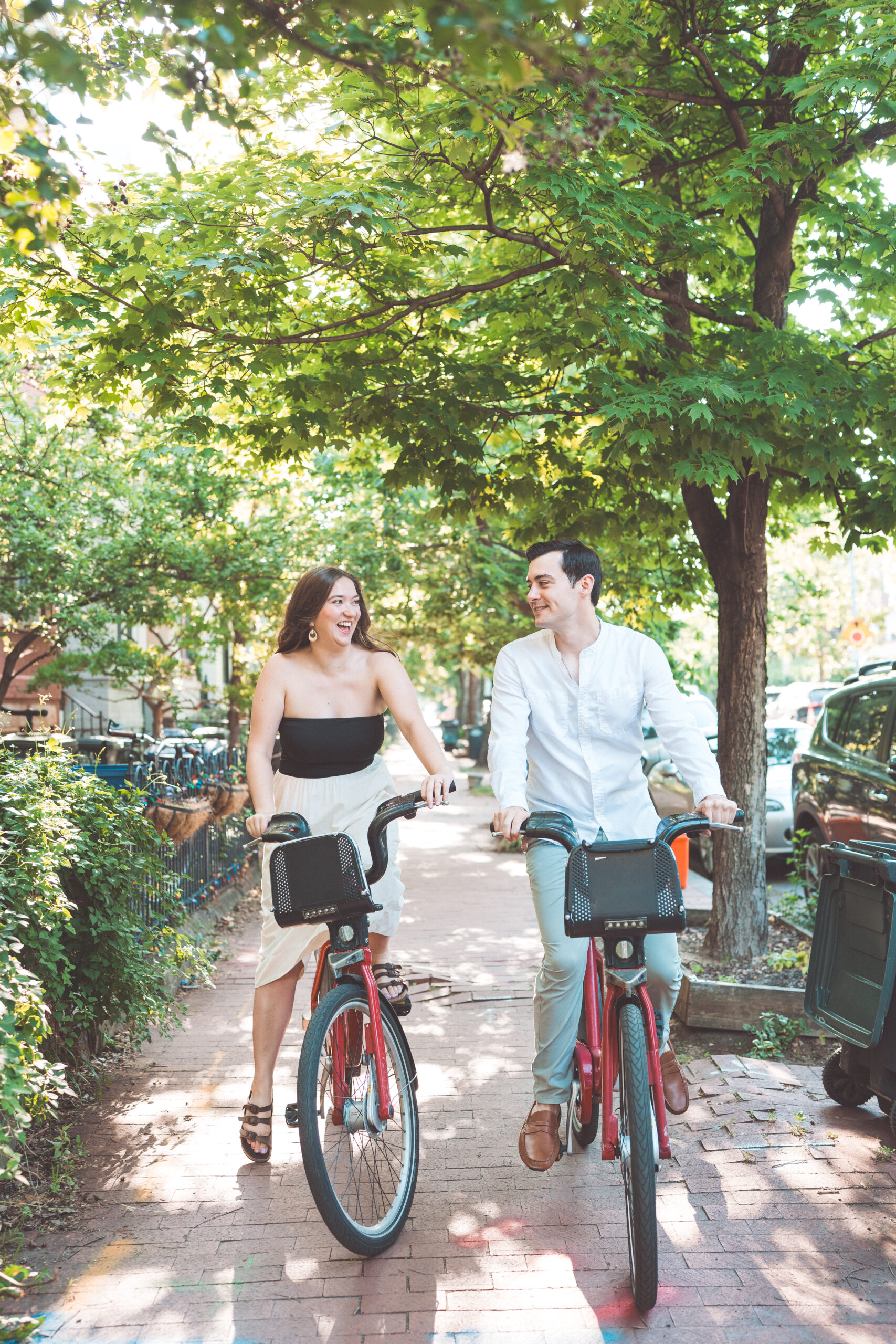 Playful Washington DC engagement photos of a couple riding Capital Bikeshare bikes down a tree-lined neighborhood street