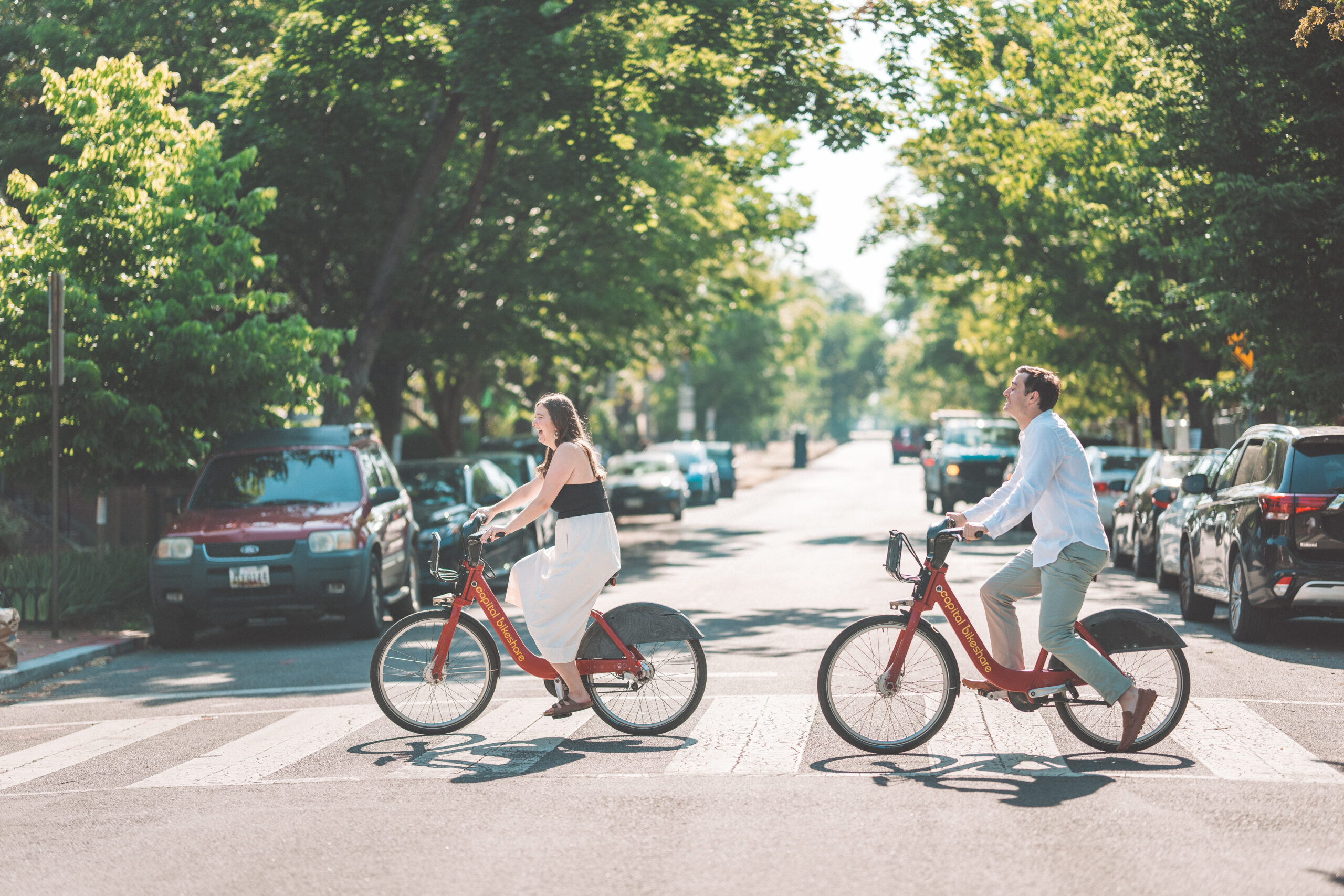 Wide shot of a couple riding red Capital Bikeshare bikes across a sunny Washington DC street during Washington DC engagement photos