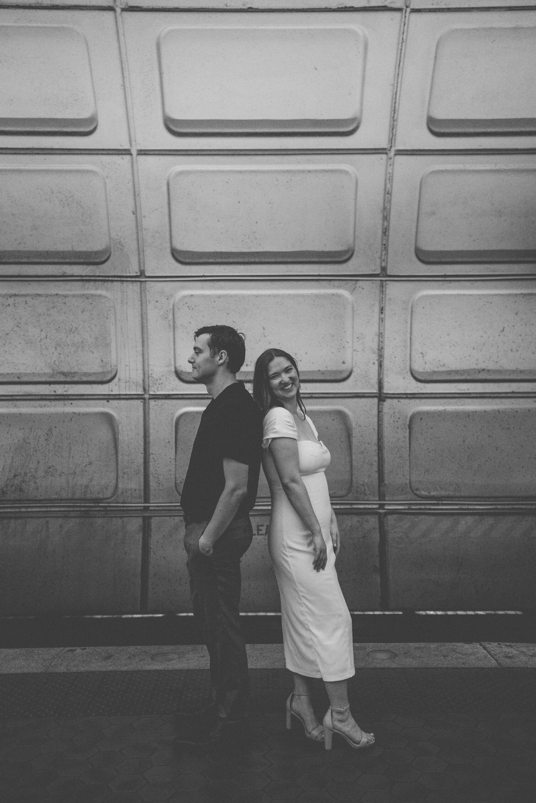 Black and white portrait of an engaged couple standing back to back against the textured metro station wall