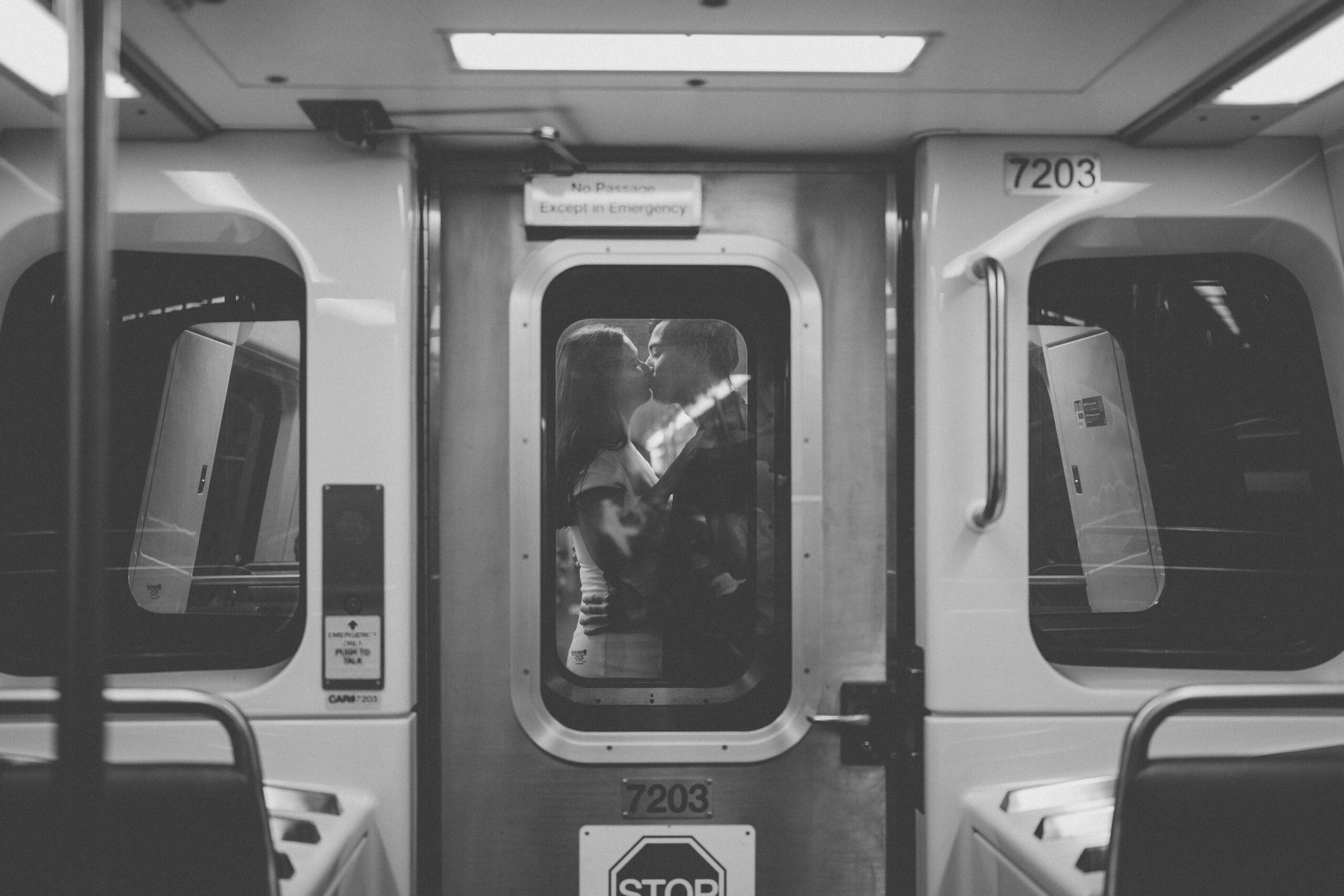 A black and white portrait of a couple sharing a kiss through the window of a subway car.