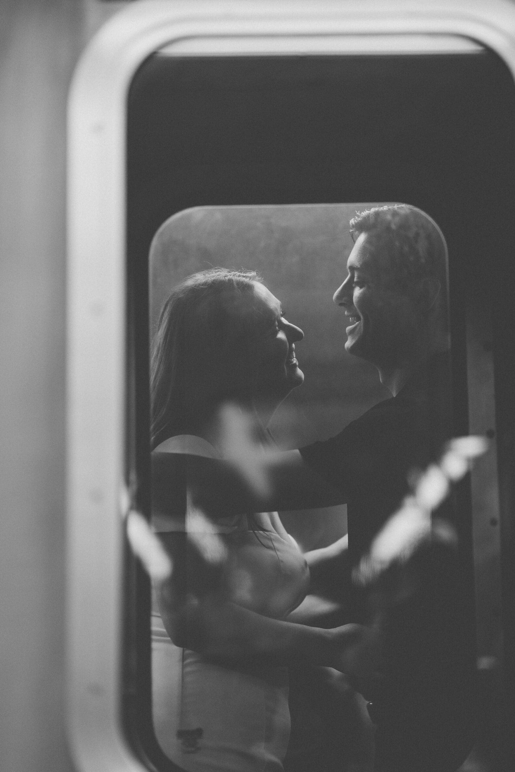 A romantic black and white portrait of a couple smiling at each other through the window of a subway car.