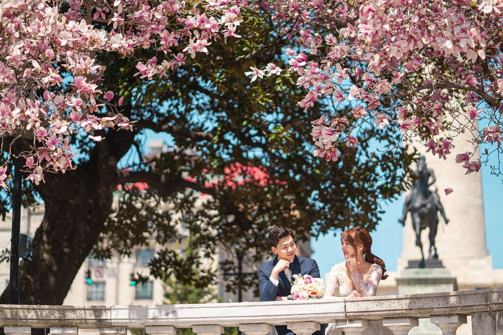 A bride and groom looking at each other while leaning on a stone railing with cherry blossoms blooming above their heads.