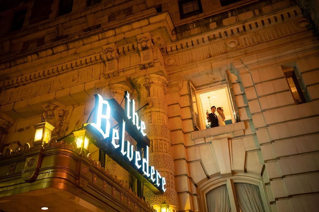 Evening portrait of a newlywed couple waving from a window above the historic Belvedere Hotel entrance.