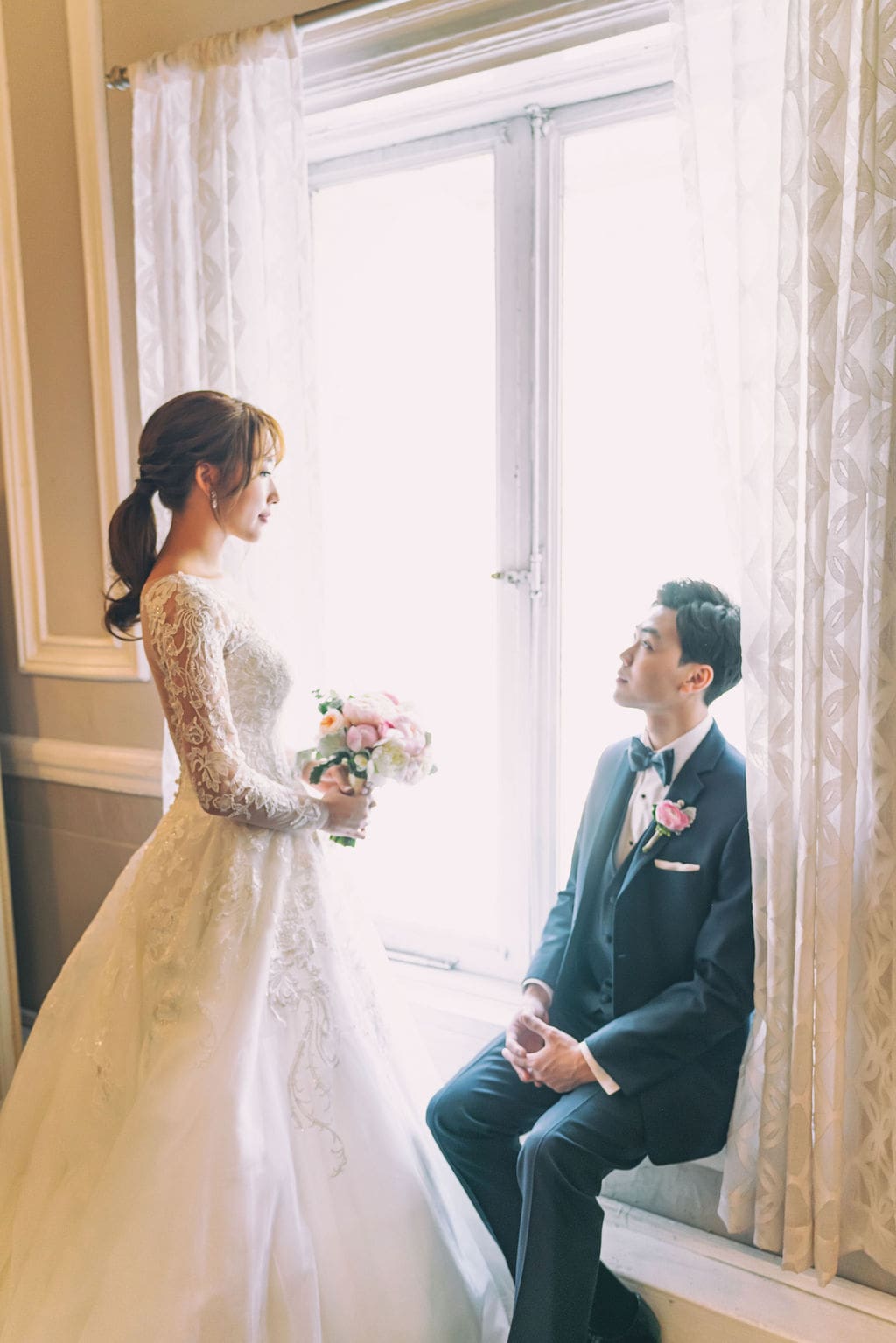 Bride and groom sharing a quiet portrait by a sunlit window before their wedding ceremony.
