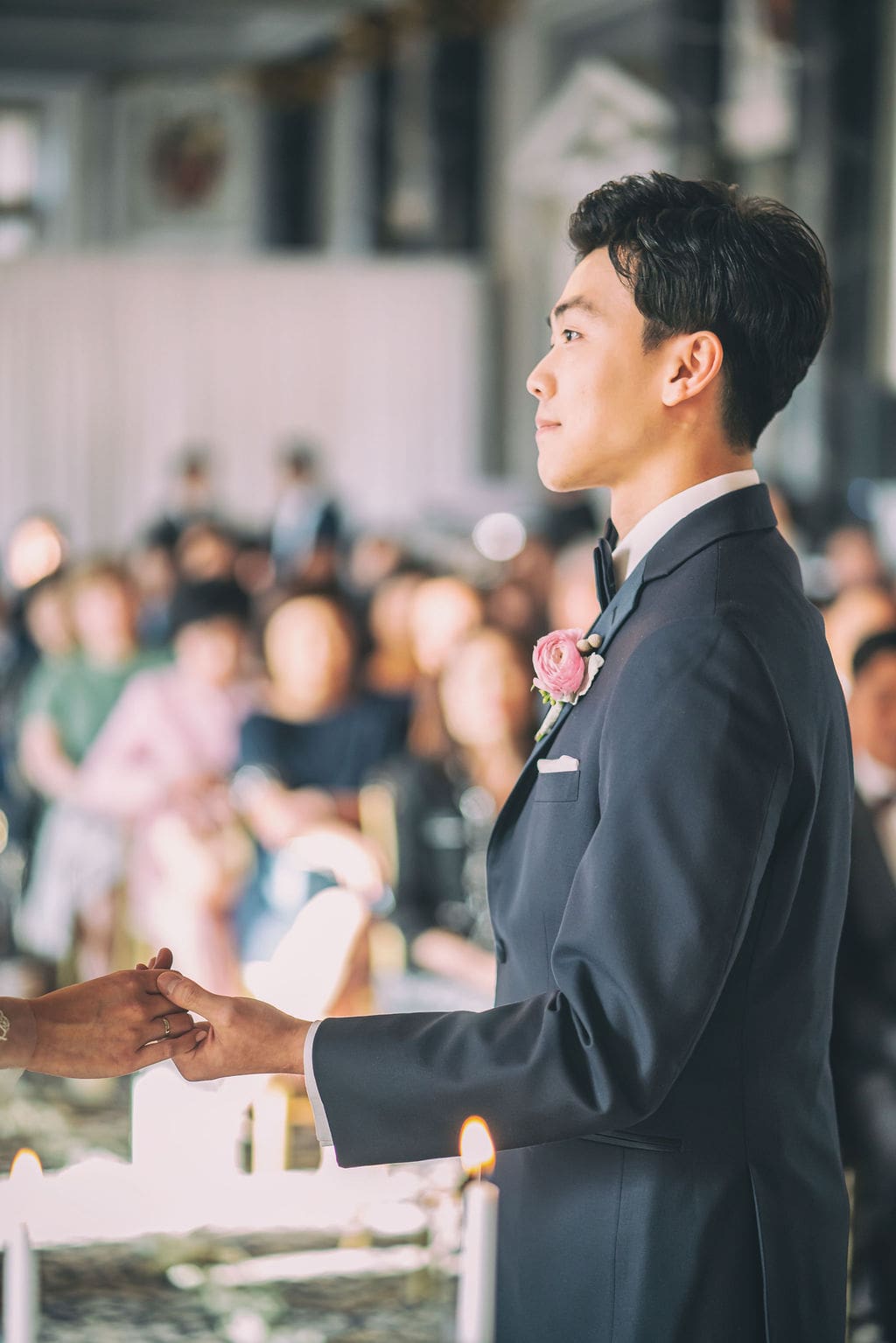 Groom holding hands with the bride during their wedding vows surrounded by family and friends.