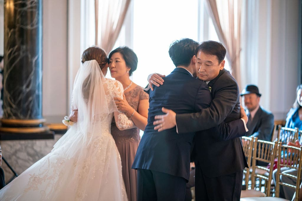 Emotional family embrace between the groom and his parents during a heartfelt wedding ceremony.