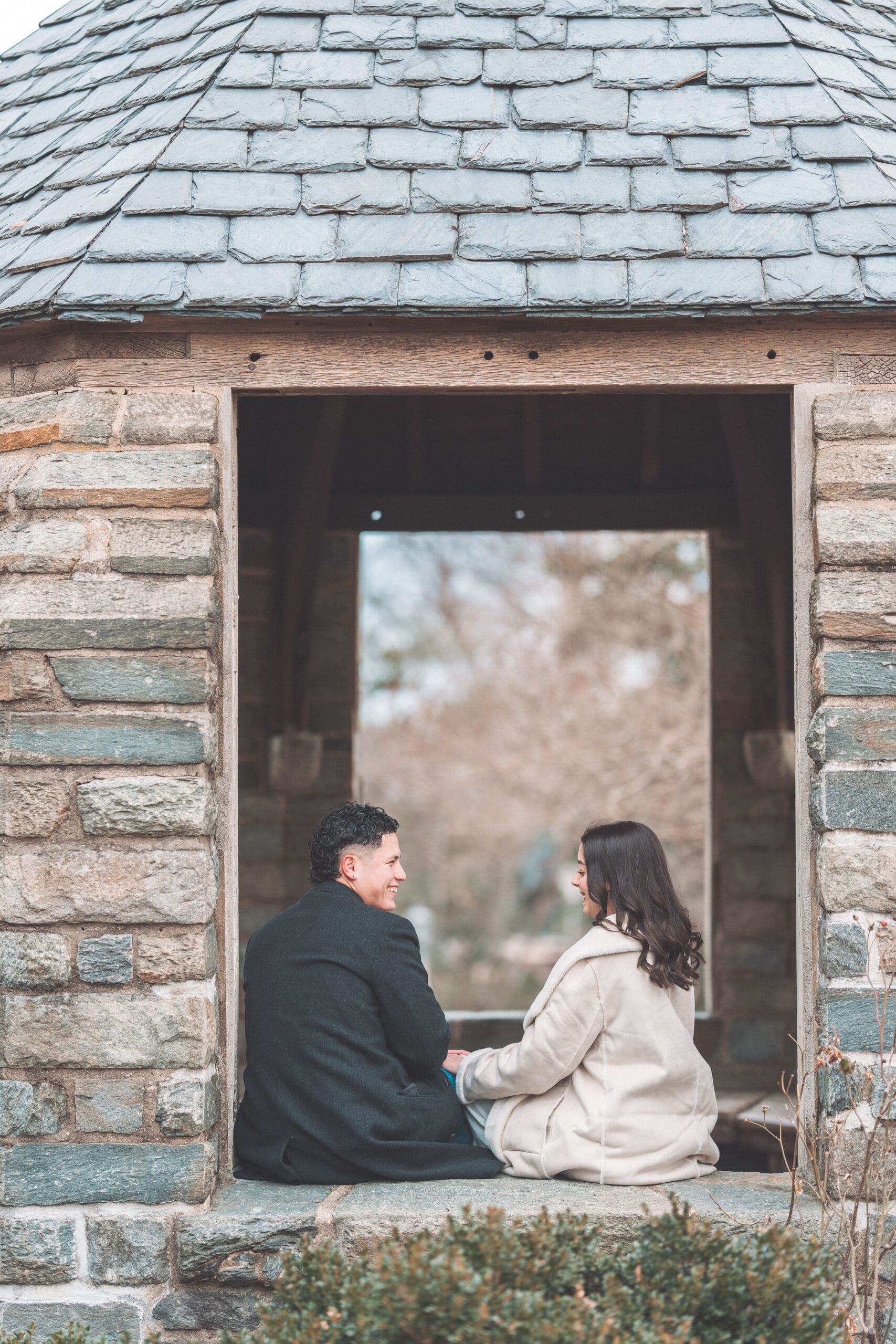 A couple sitting next to each other in the window of a stone cathedral, laughing at each other and holding hands.