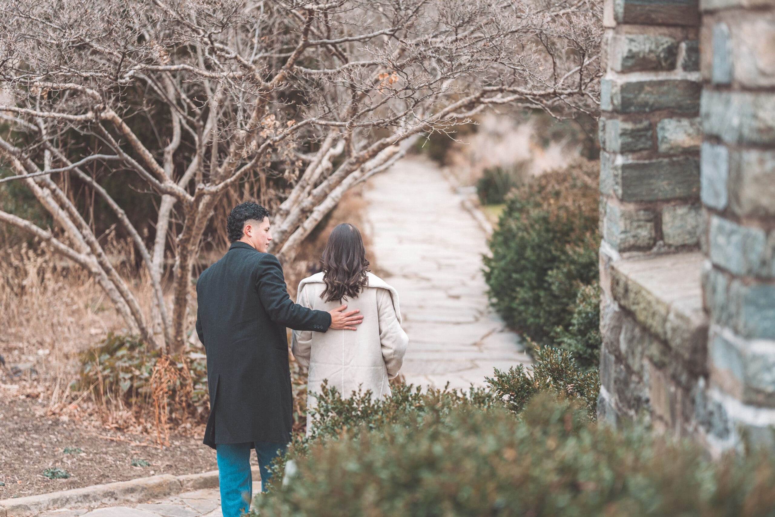 A newly engaged couple walk down a stone path side by side with their backs to the camera.