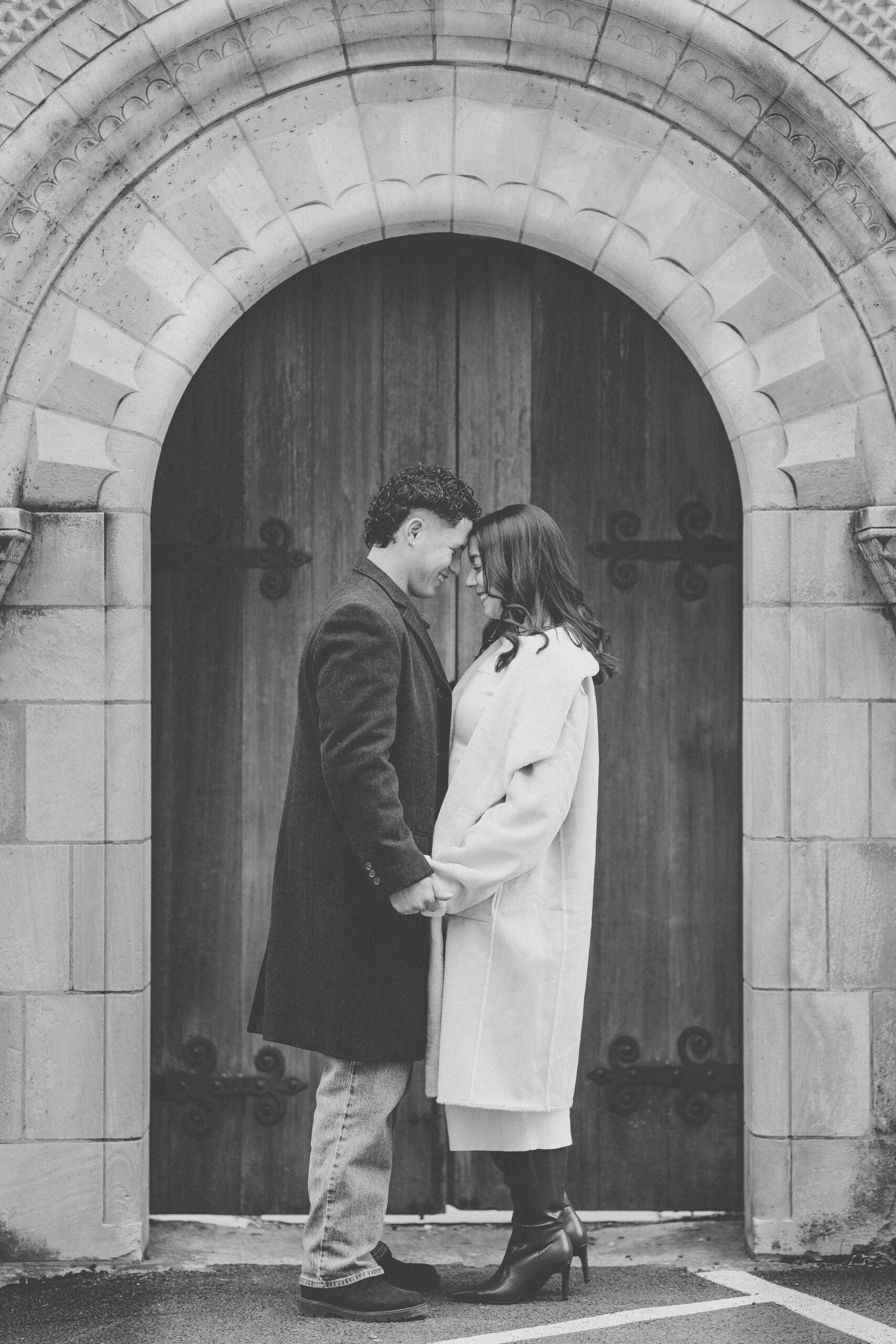 Black and white photo of a newly engaged couple sharing a quiet moment in front of the cathedral’s grand wooden doors during their portrait session.