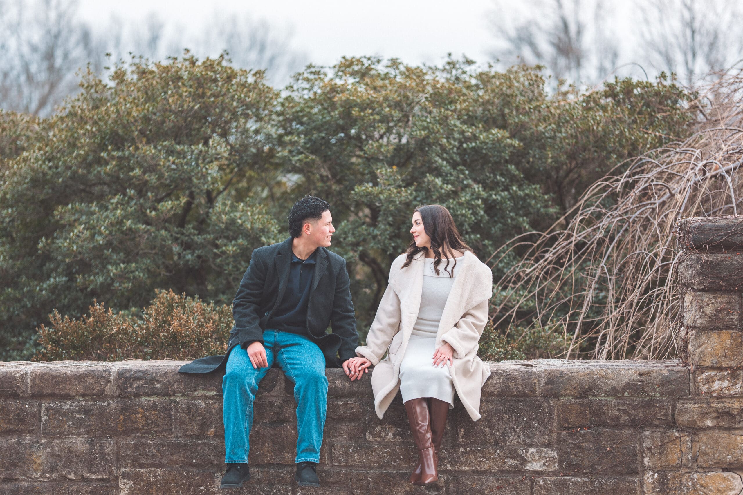 A couple sitting on a stone ledge looking at each other lovingly.