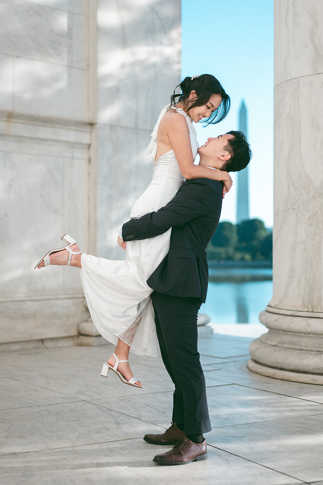 Groom lifting the bride beneath the Jefferson Memorial with the Washington Monument in the background during a Washington DC engagement session.
