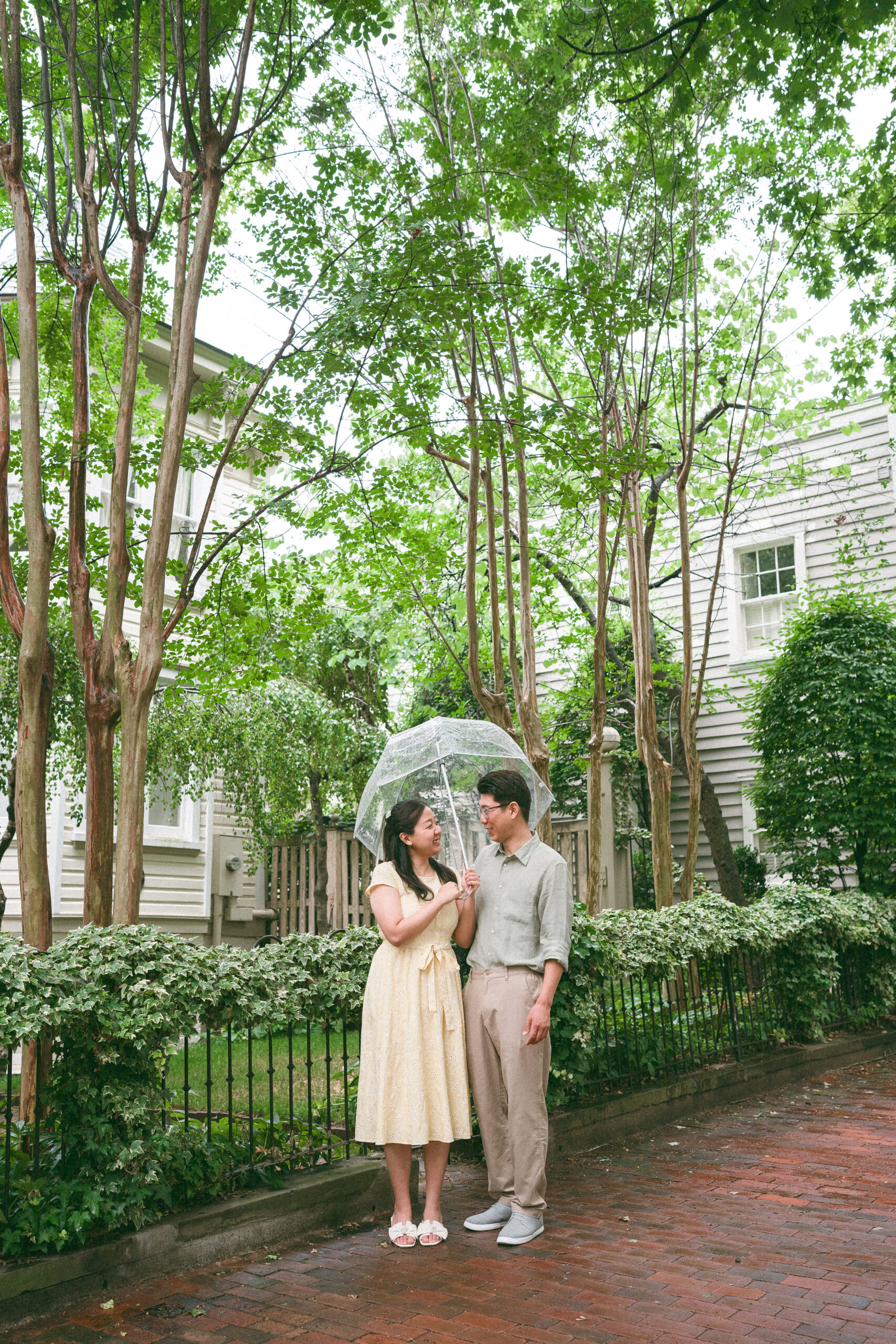 Couple standing beneath a clear umbrella on a tree-lined neighborhood street during rainy engagement photos.