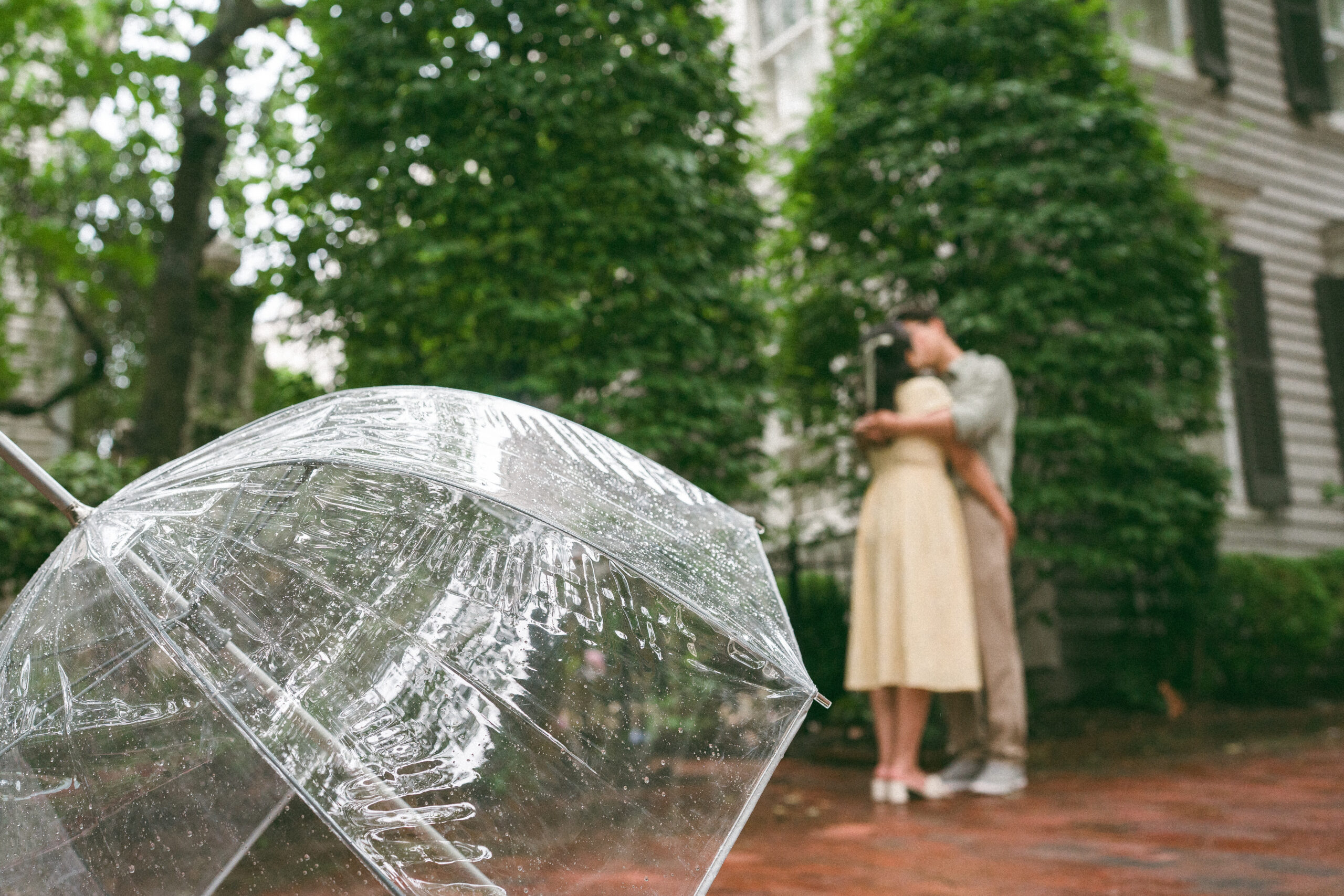 Close-up of a rain-covered clear umbrella with the couple embracing softly in the background.