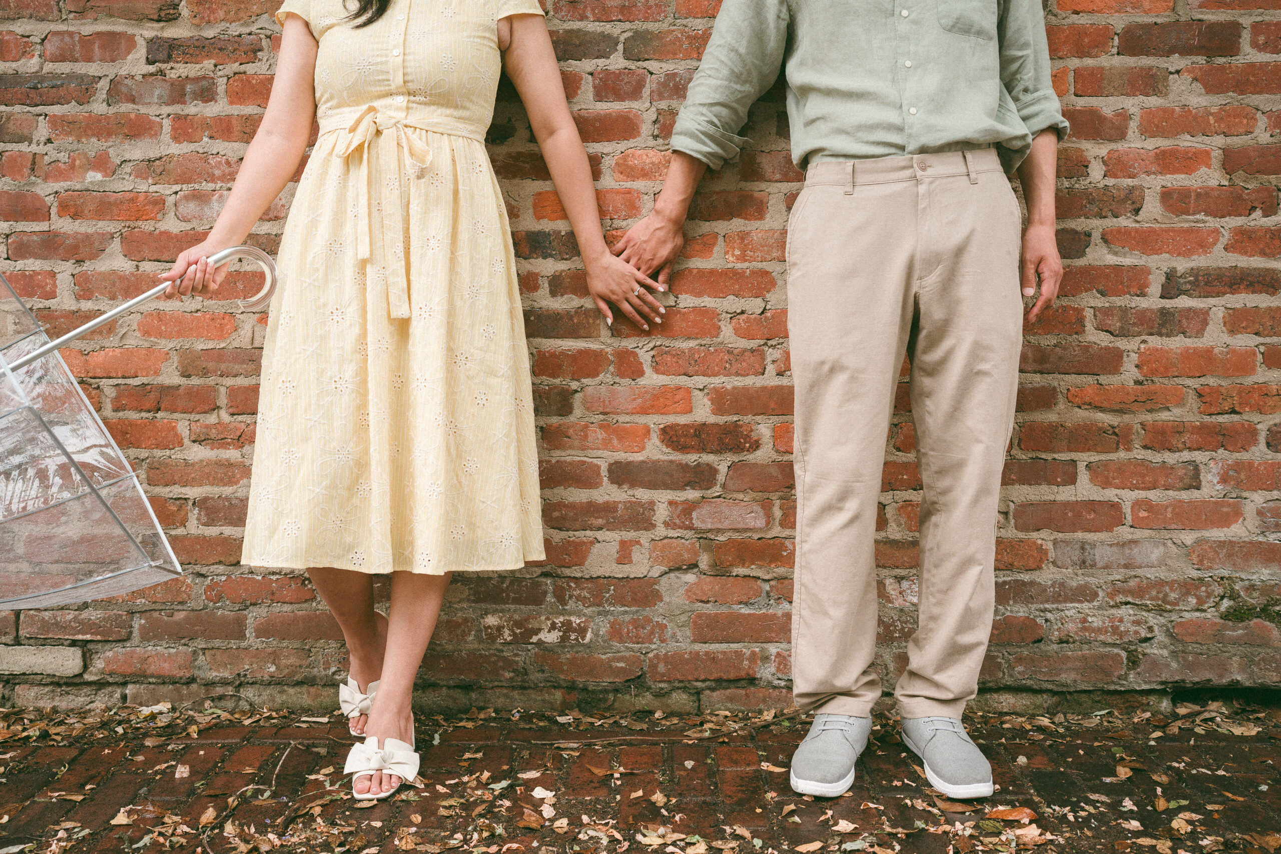 Close-up of the couple holding hands against a brick wall with a clear umbrella nearby.