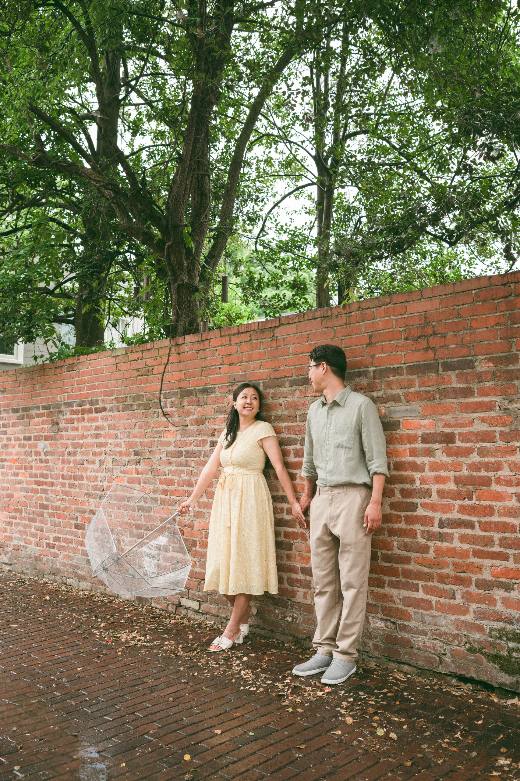 Couple holding hands and smiling beside a historic brick wall after the rain.