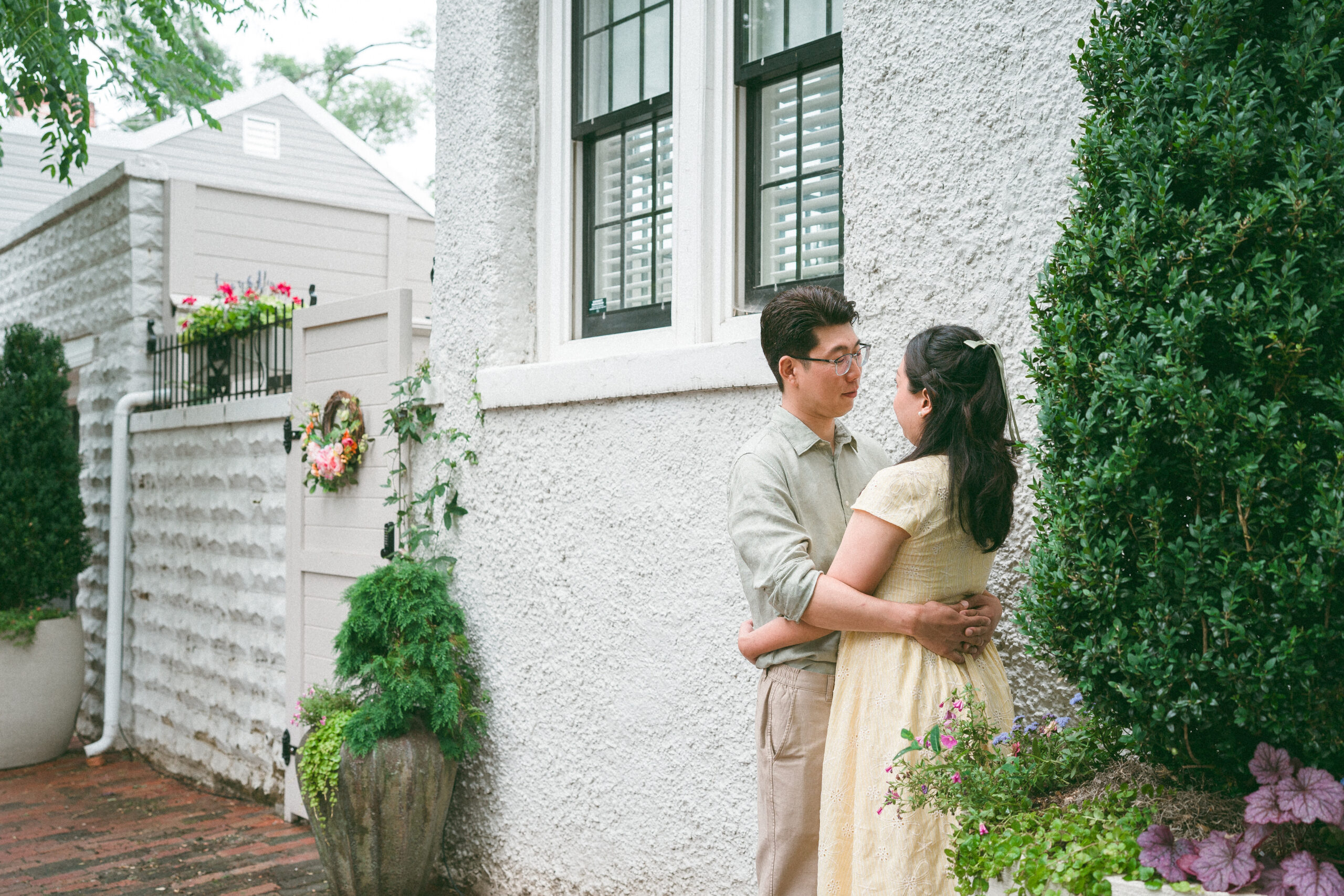 Couple embracing beside a textured white wall surrounded by greenery during rainy engagement photos.
