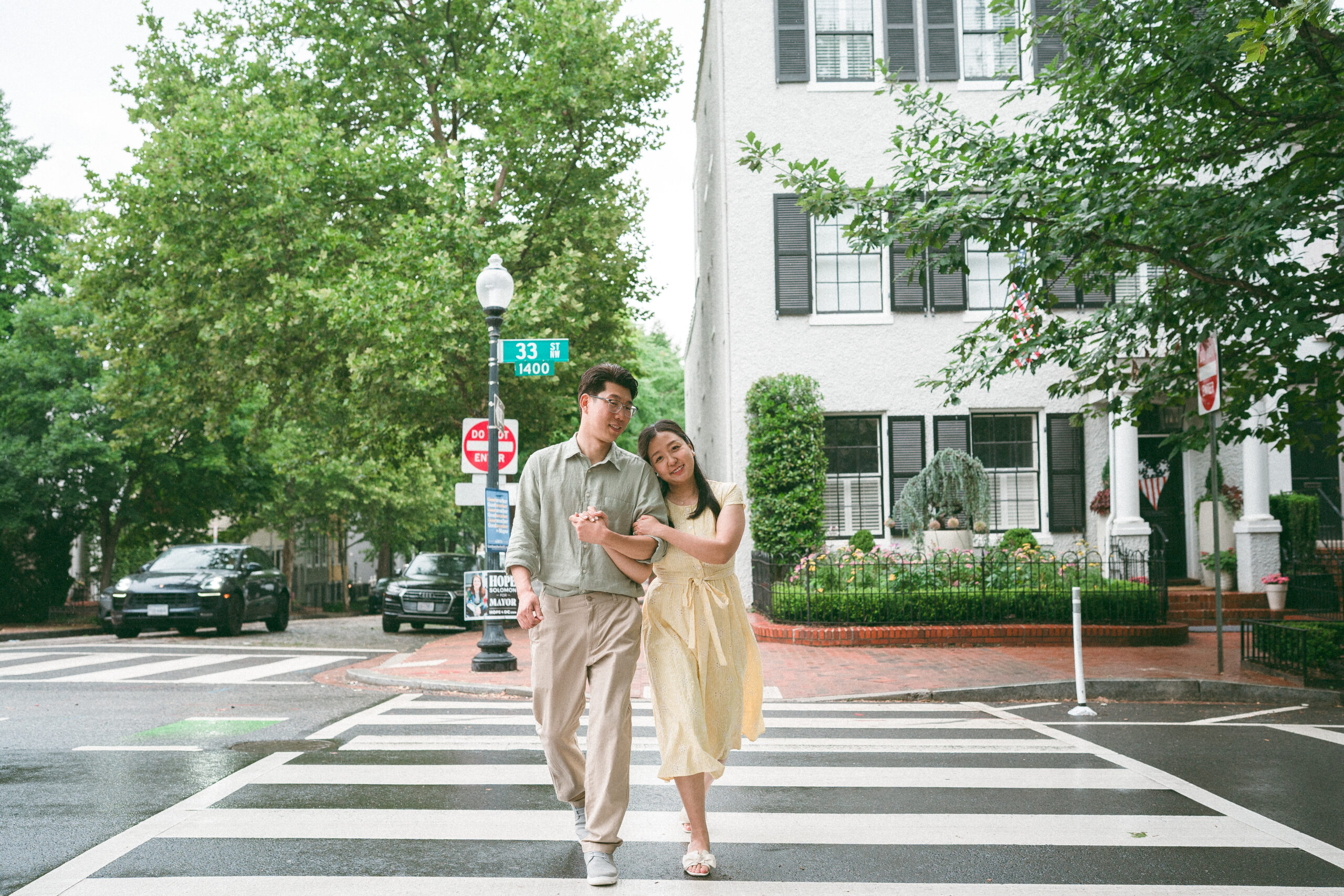 Couple walking arm in arm across a neighborhood crosswalk, smiling as they enjoy the rainy day.