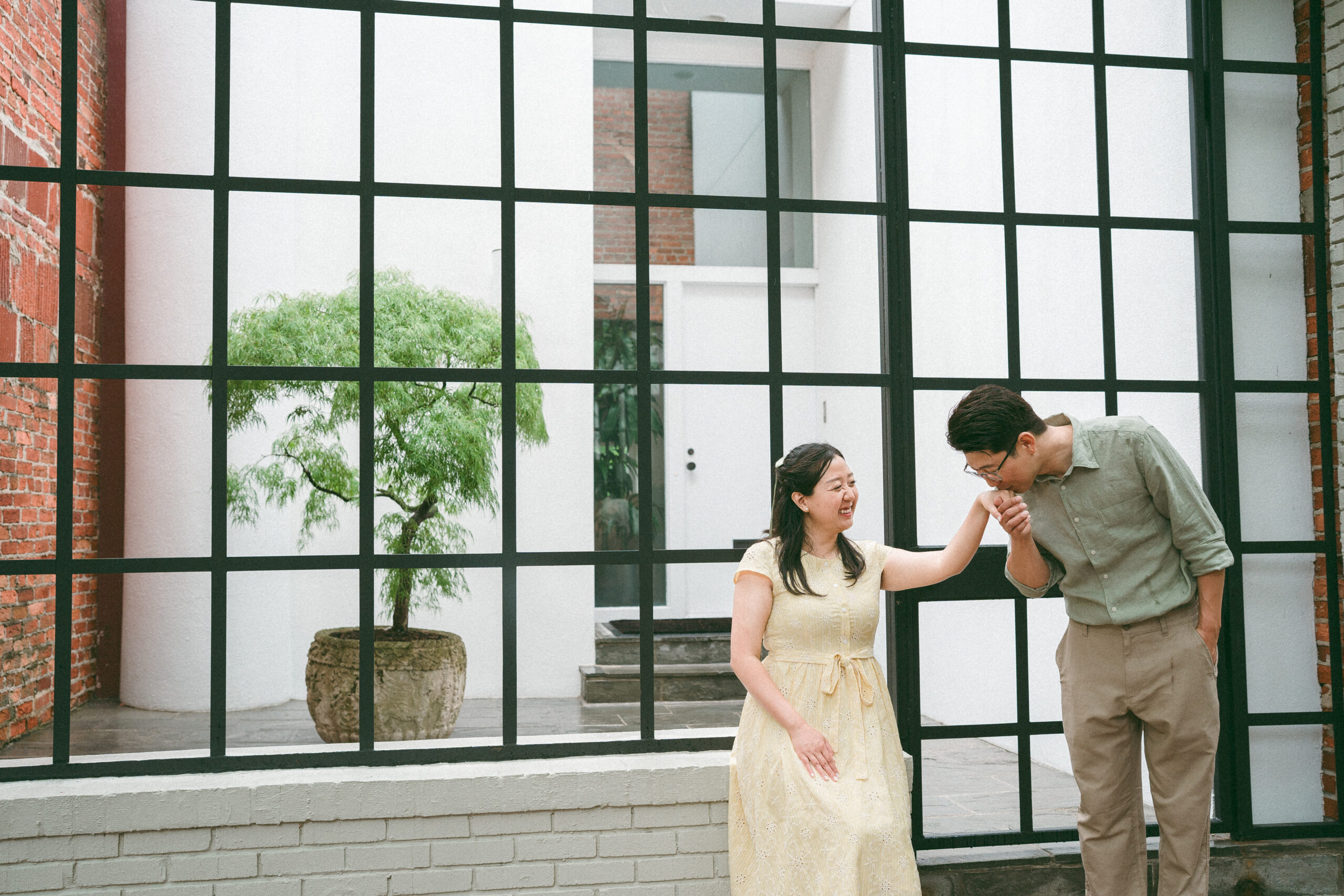 Groom kissing his partner's hand in front of a modern black-framed building during rainy engagement photos.