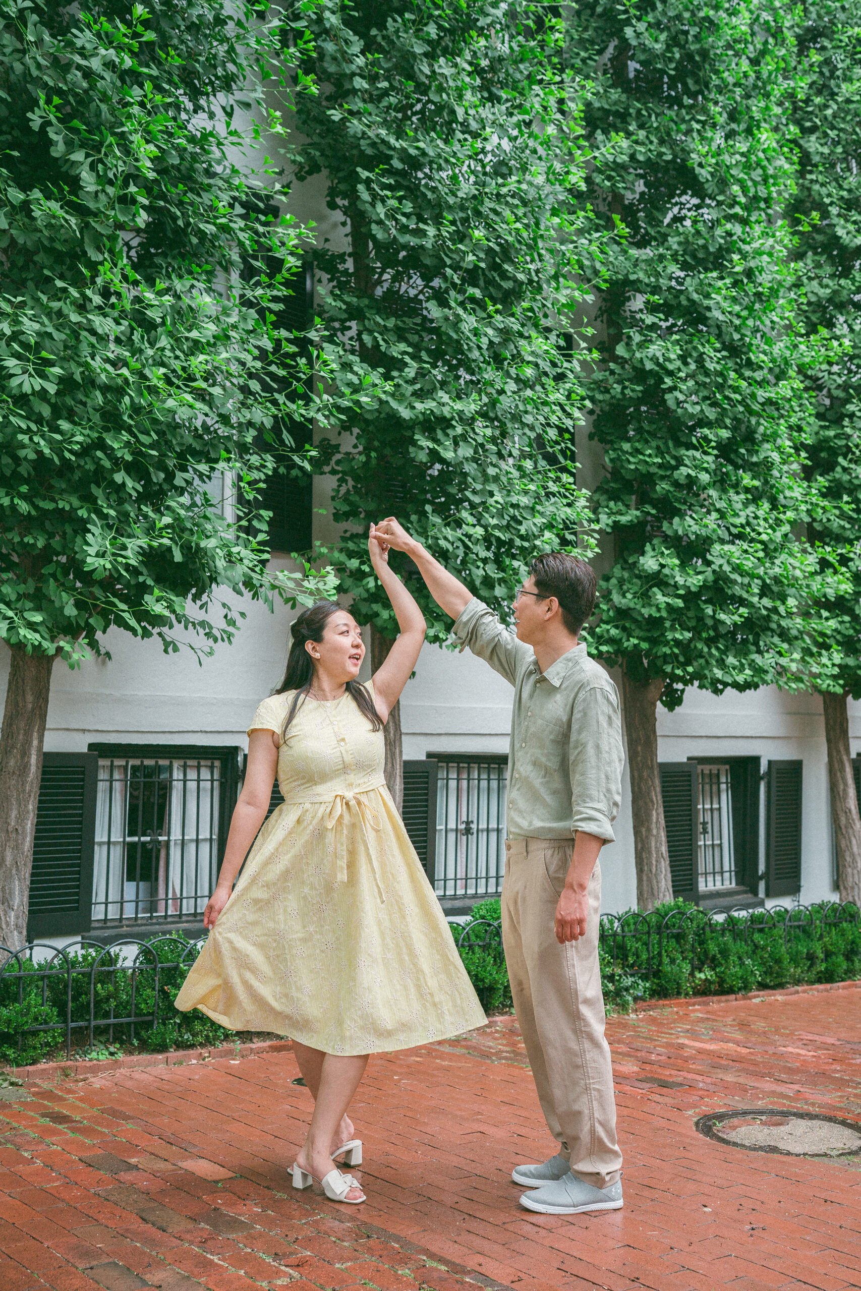 Couple twirling together beneath tall green trees during rainy engagement photos.