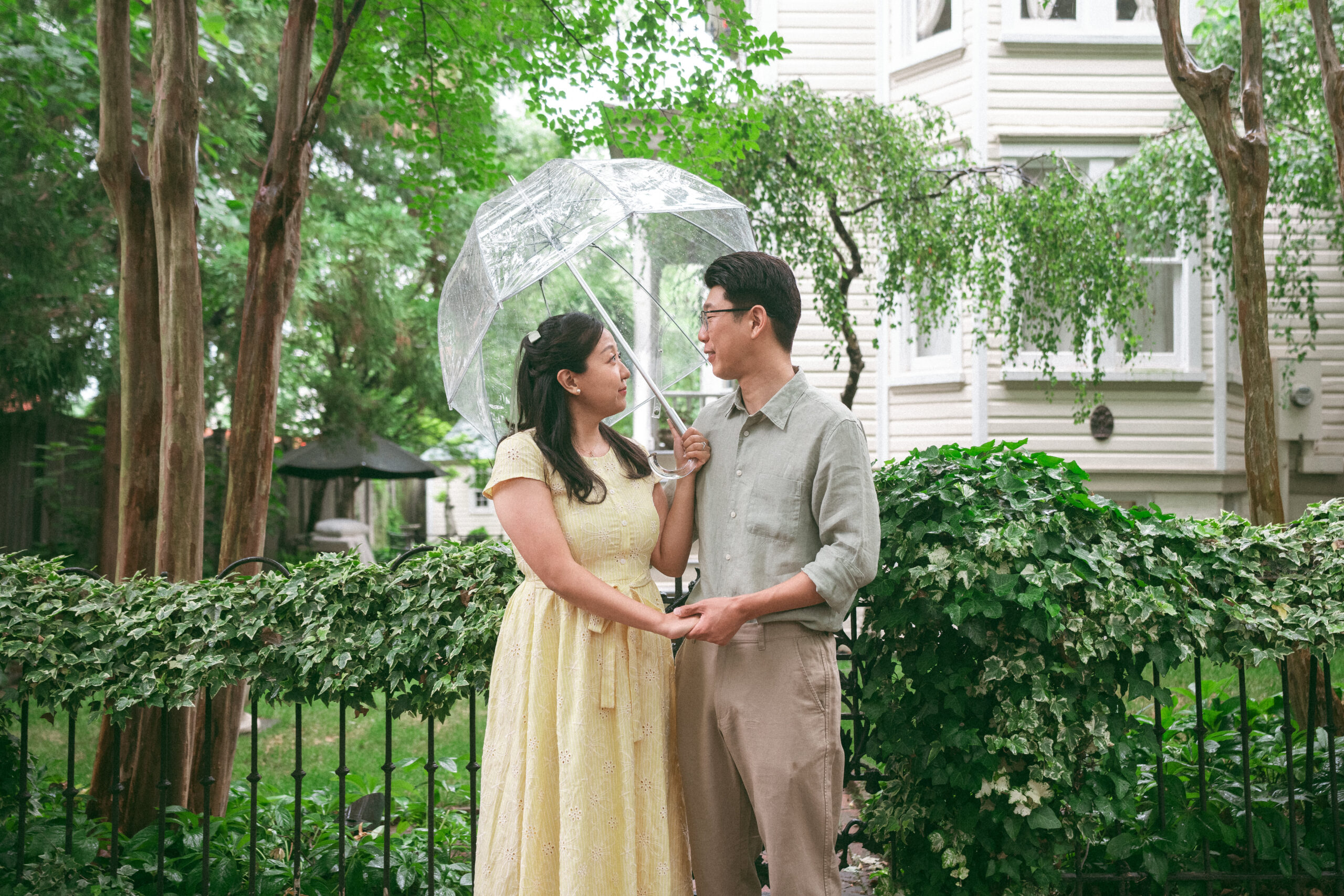 Couple standing beneath a clear umbrella and holding hands in a lush garden setting.