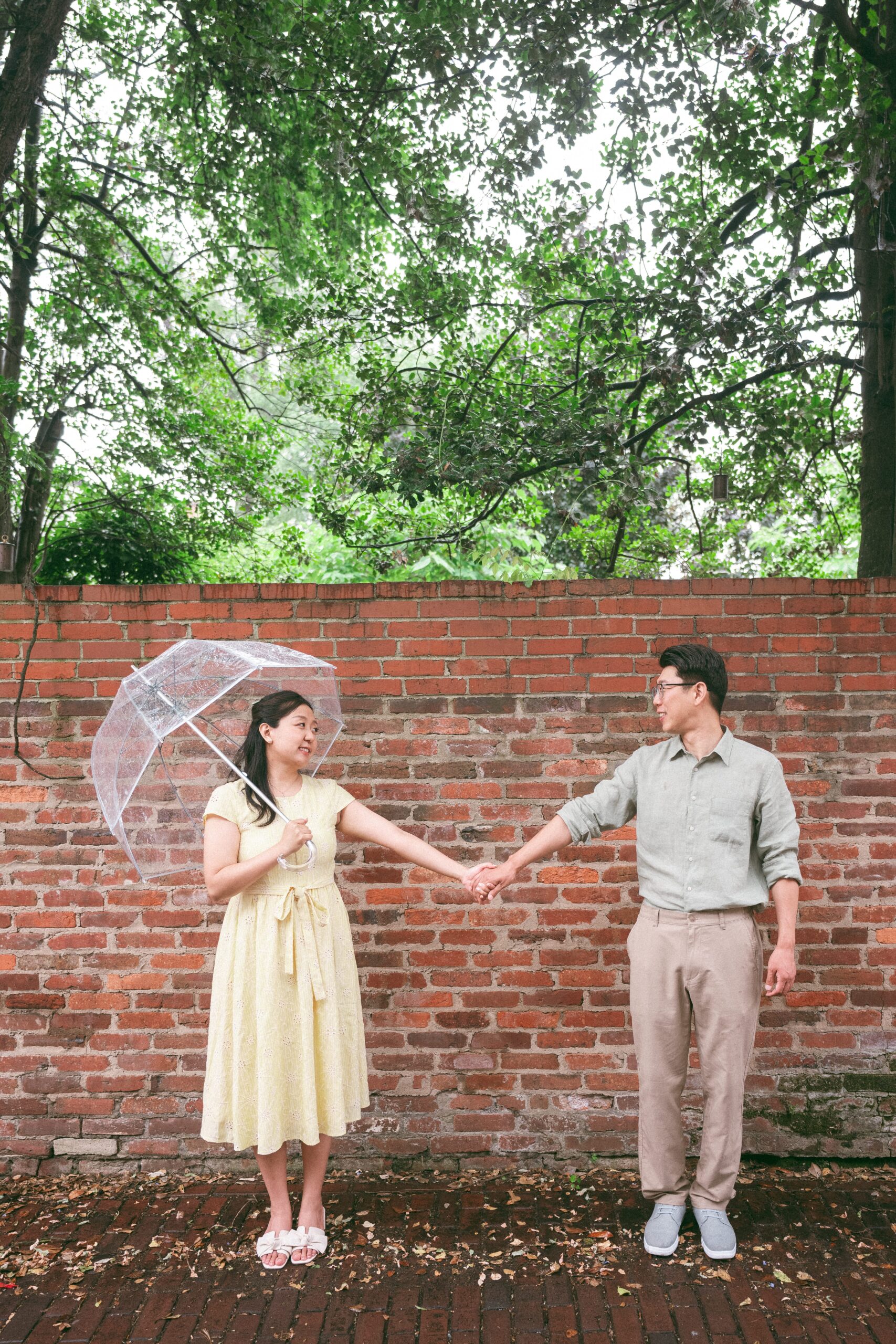 Couple holding hands in front of a brick wall with a clear umbrella during a rainy engagement photos session.