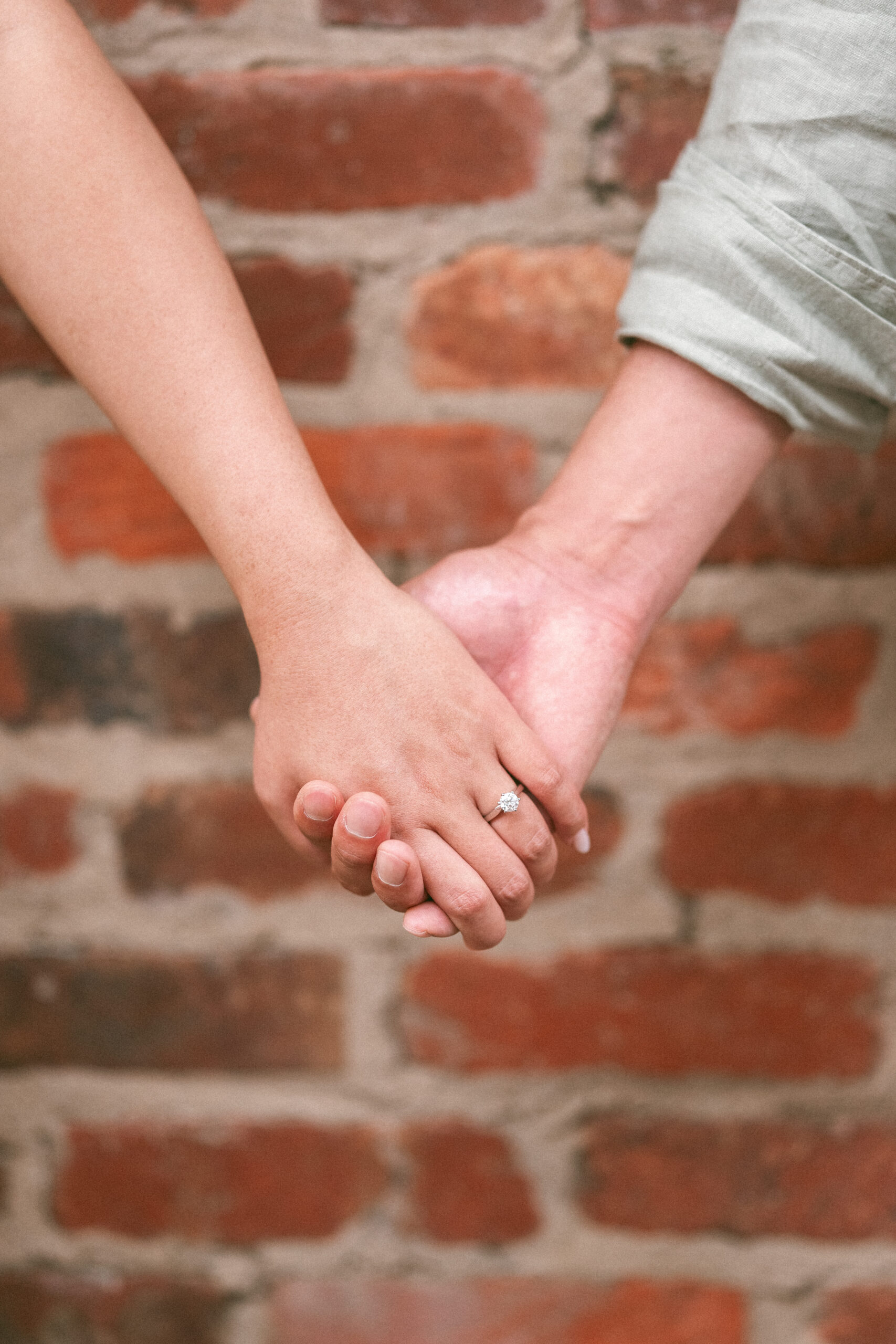 Close-up of the couple holding hands with an engagement ring in focus against a brick wall.