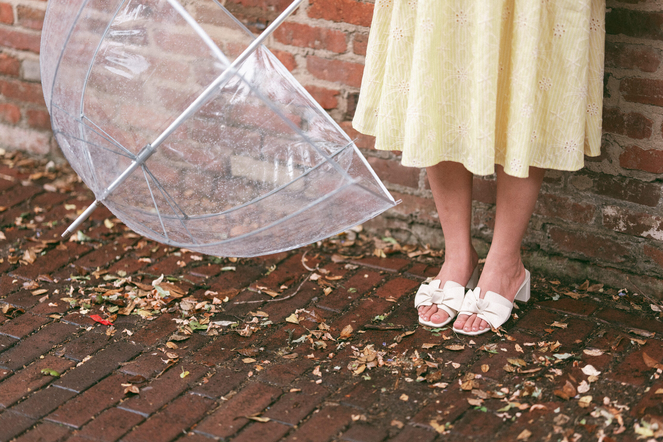 Detail photo of a clear umbrella resting beside the bride-to-be's shoes on a brick sidewalk during rainy engagement photos.