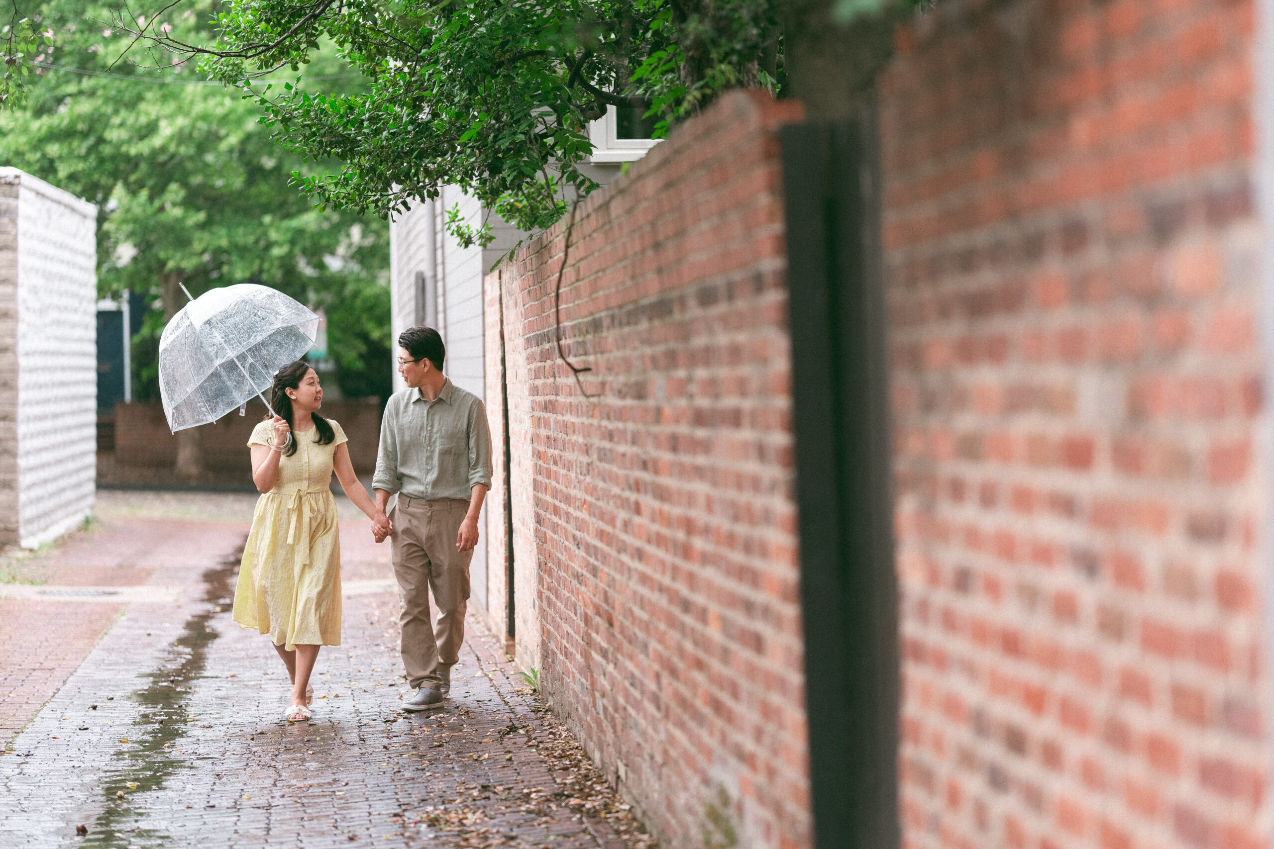 Couple strolling hand in hand beneath a clear umbrella along a quiet brick alley.