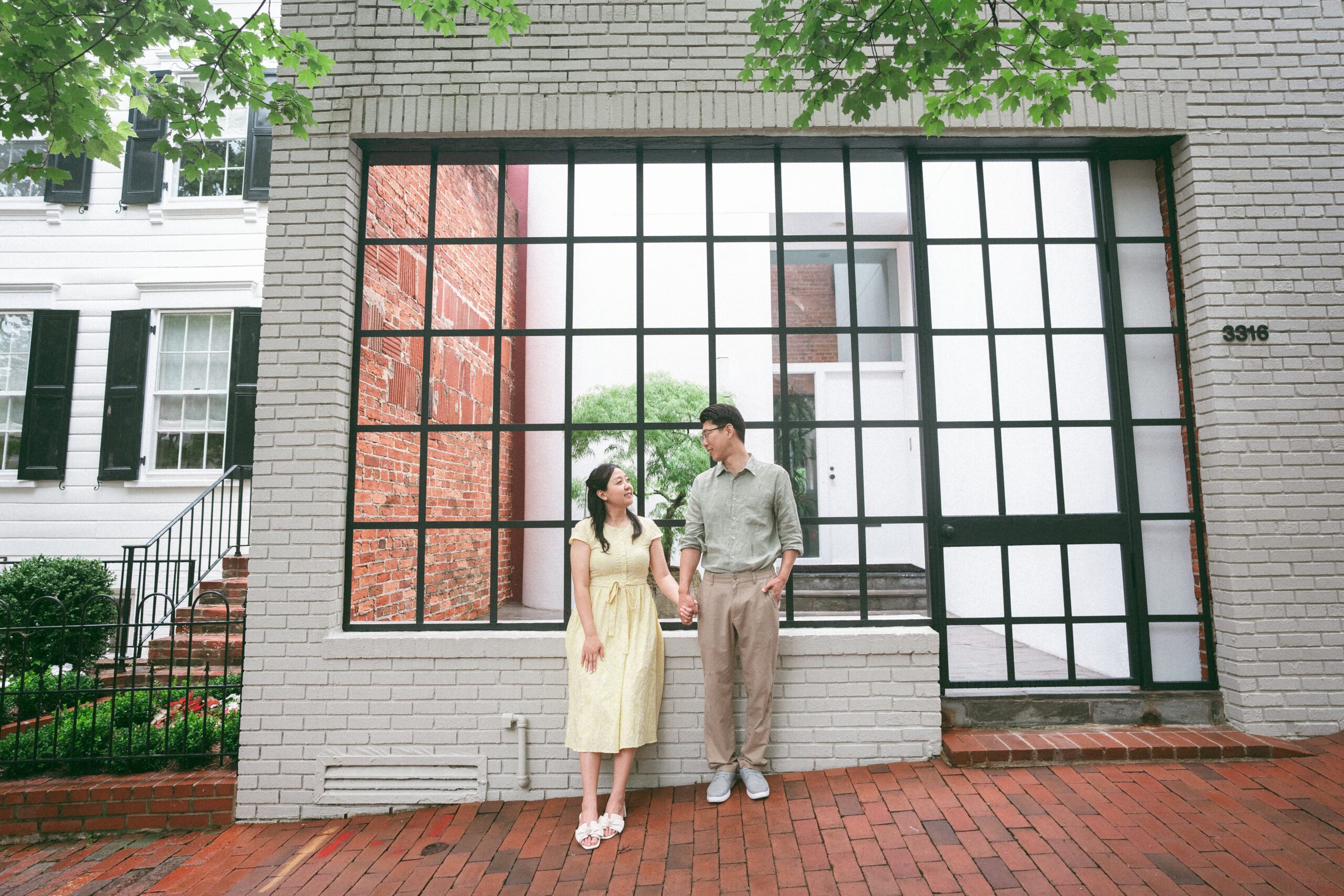 Couple holding hands in front of a modern black-framed window during a rainy engagement photos session.