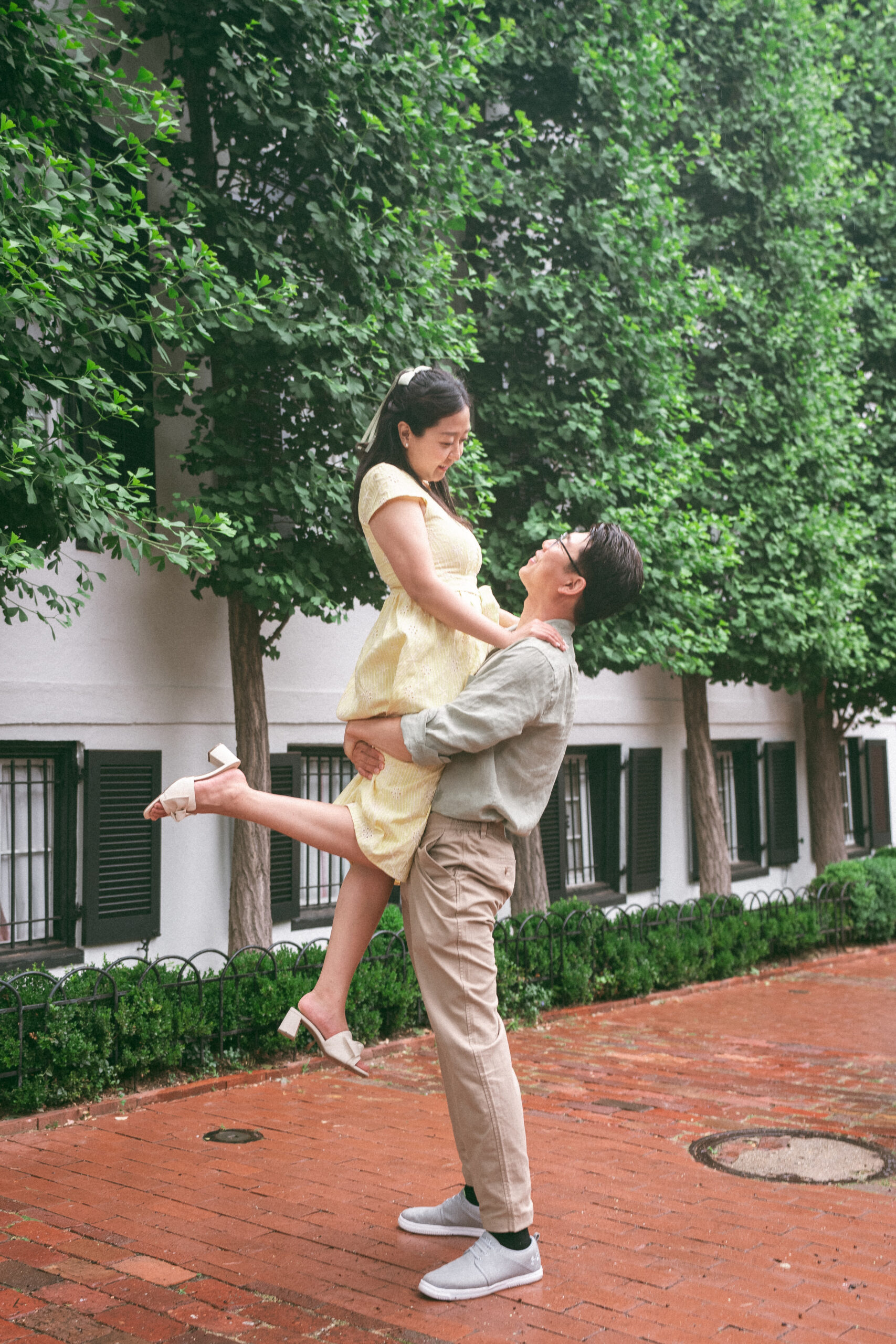 Groom lifting his partner into the air as they smile together on a brick sidewalk during rainy engagement photos.