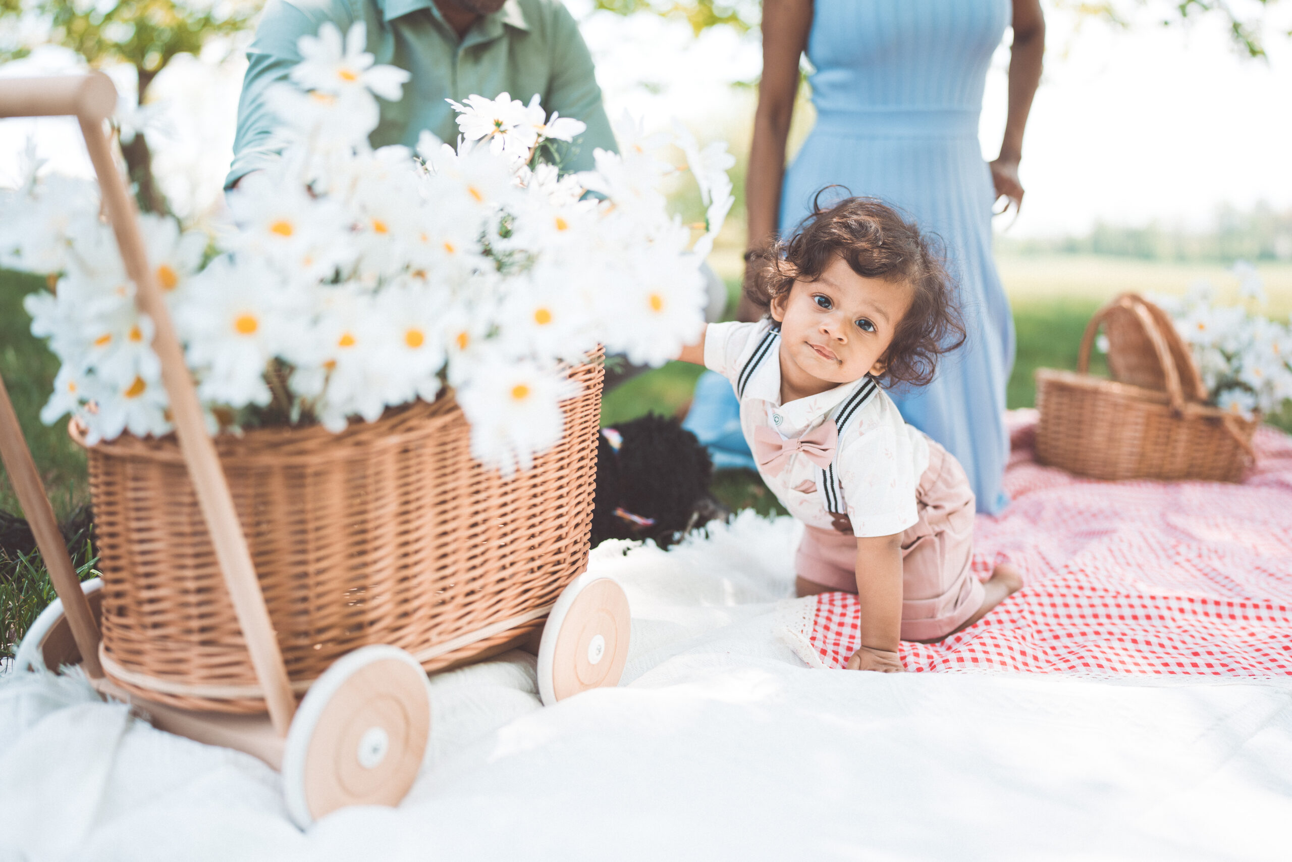 Toddler crawling beside a wicker cart filled with white daisies during an outdoor family session.