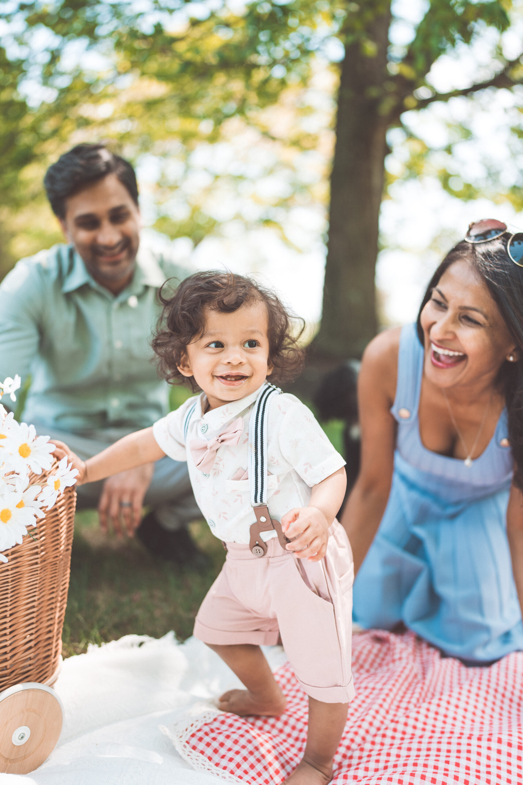 Toddler smiling while standing on a picnic blanket as parents laugh together during a Maryland family photoshoot in the park.
