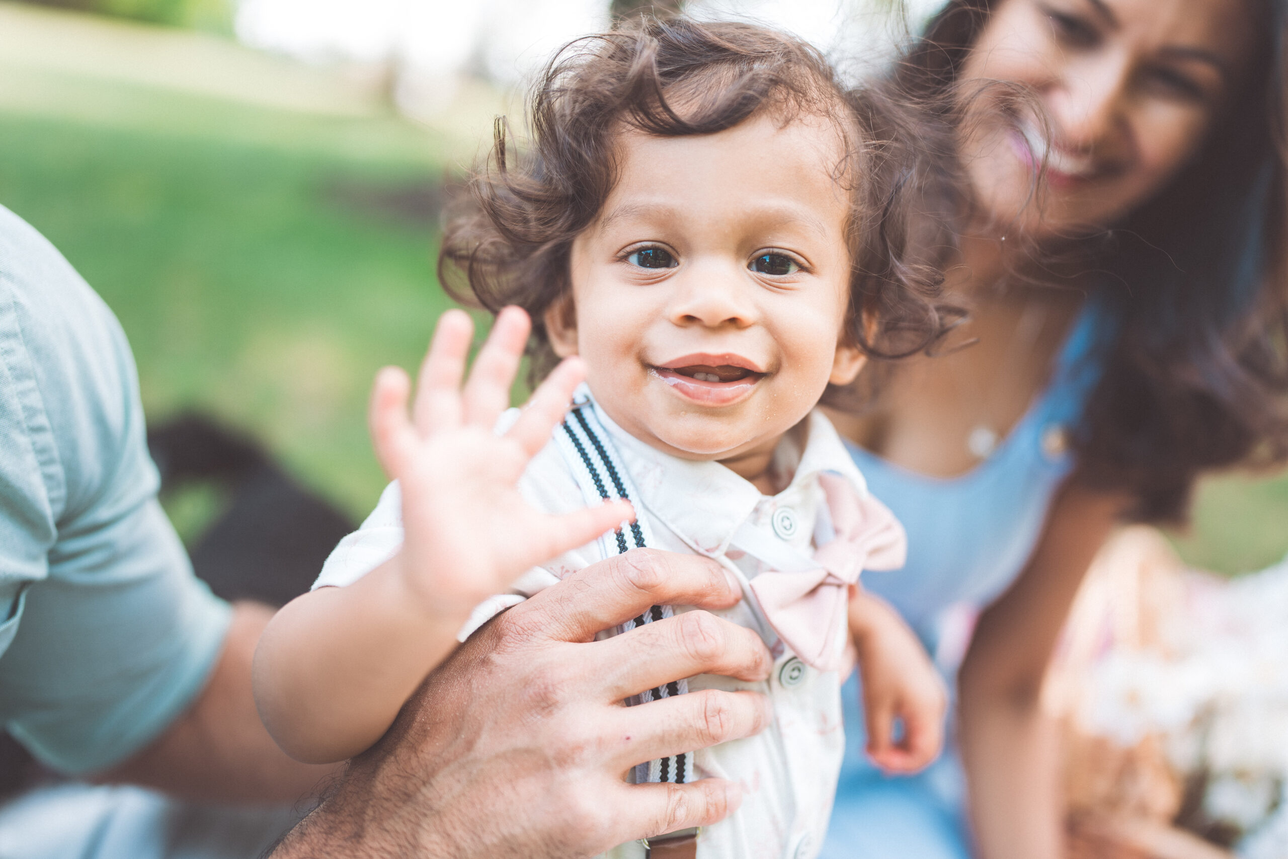 Toddler waving at the camera while being held by a parent during an outdoor family session.