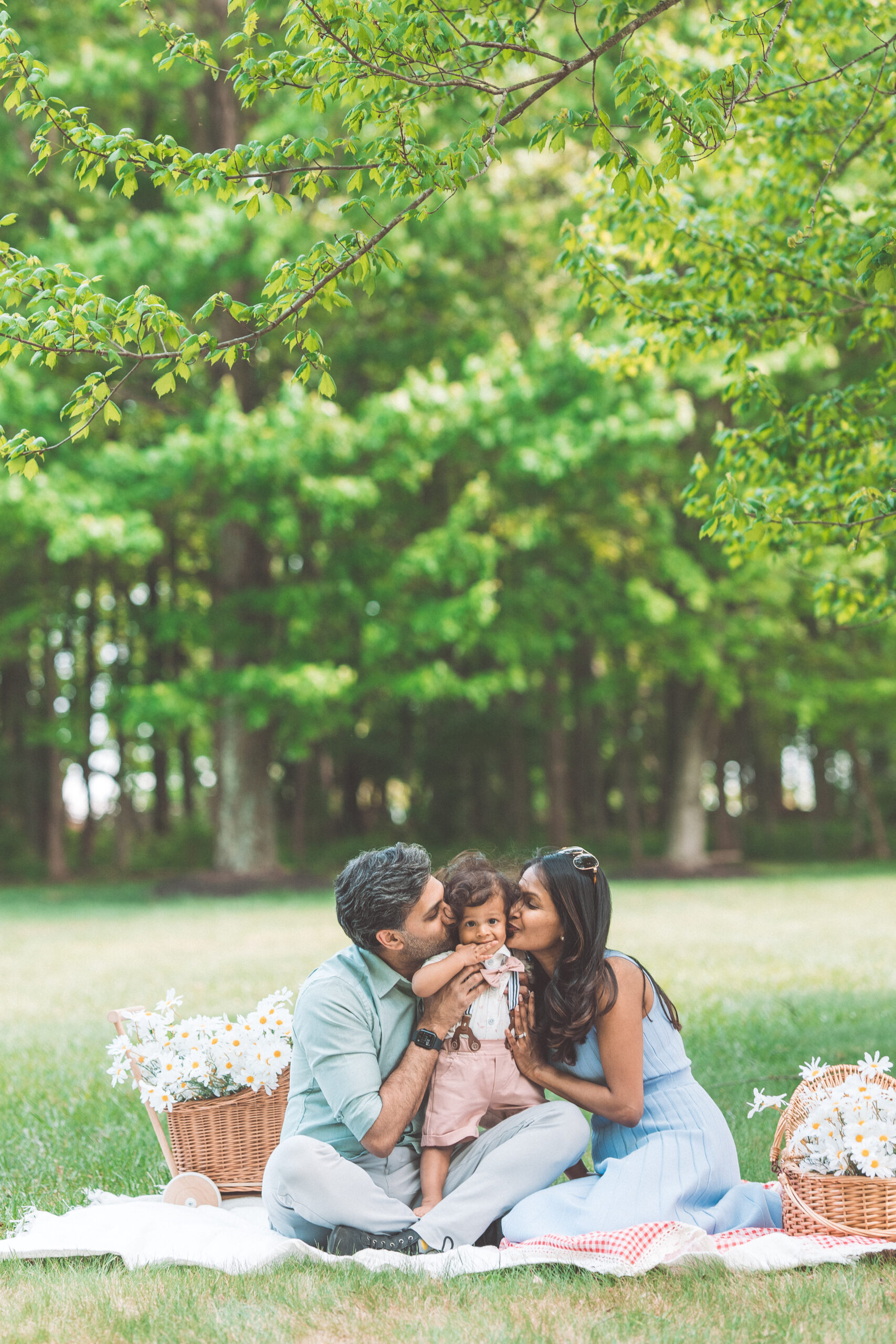 Parents kissing their smiling toddler on both cheeks during a playful picnic session.