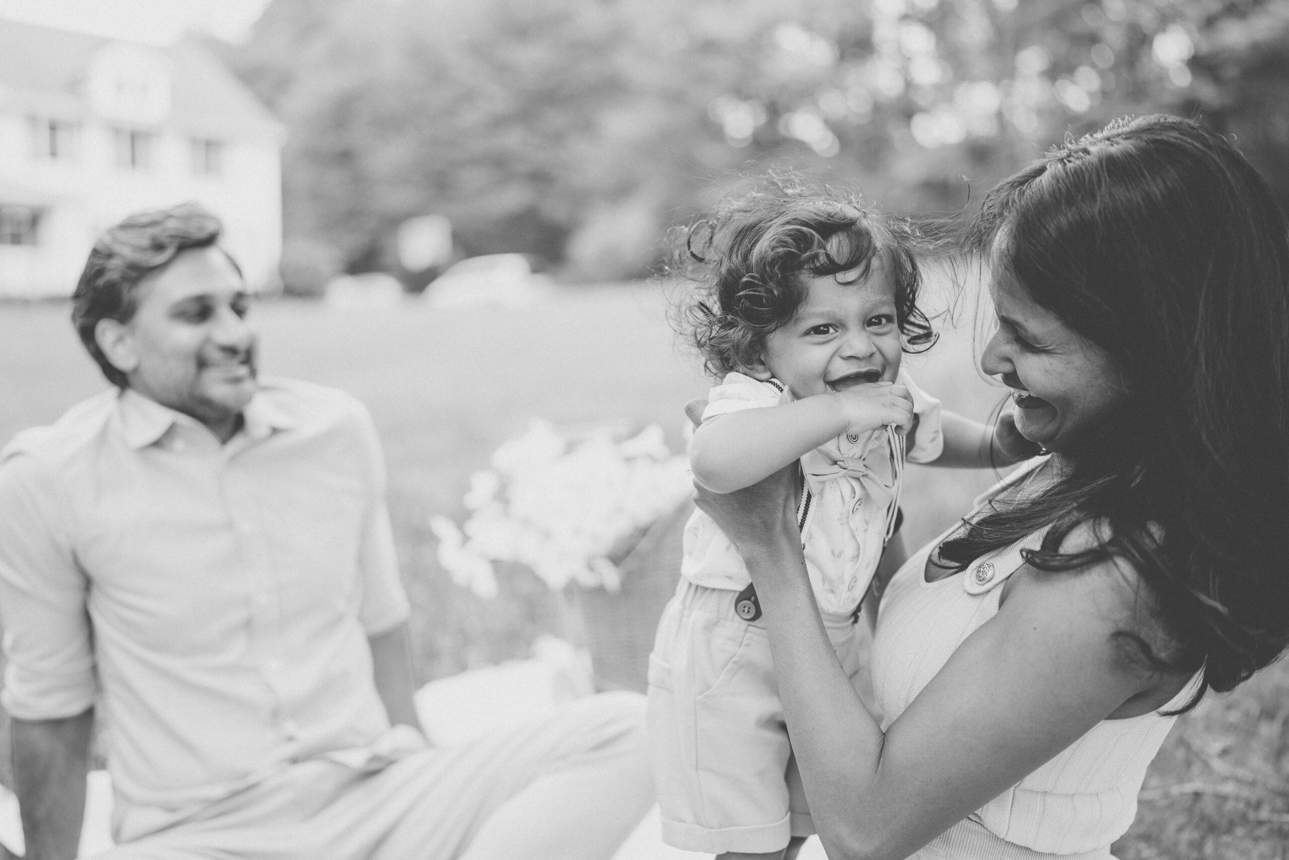 Black and white candid of a mother holding her laughing toddler while dad smiles in the background.