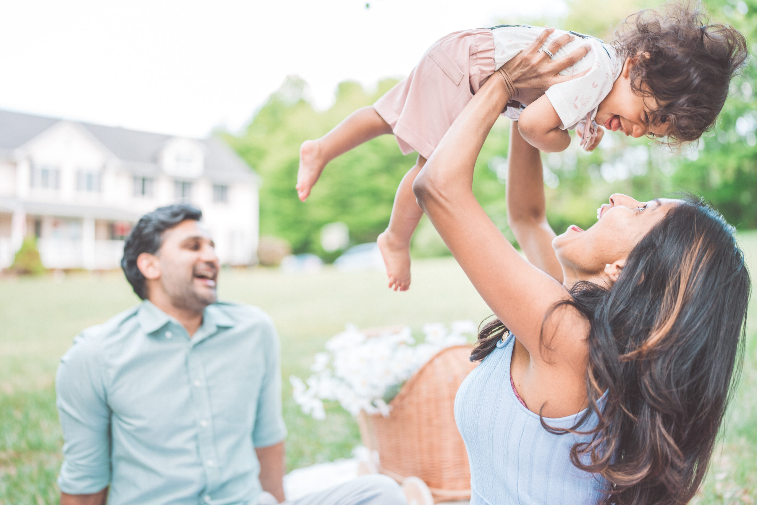 Mother lifting her toddler into the air while dad smiles in the background during an outdoor family session.