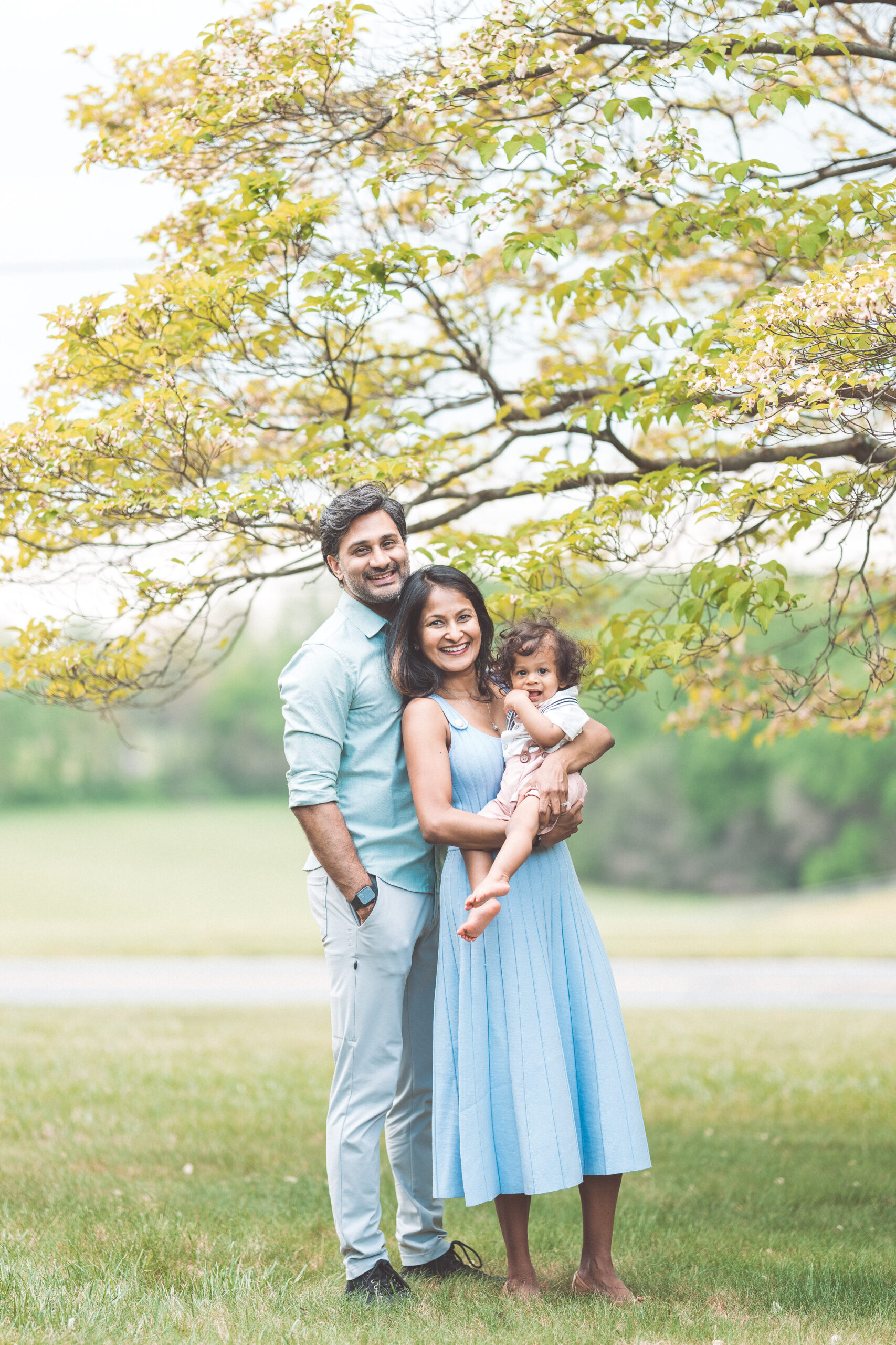 Family of three standing together beneath a leafy tree in a relaxed outdoor portrait.