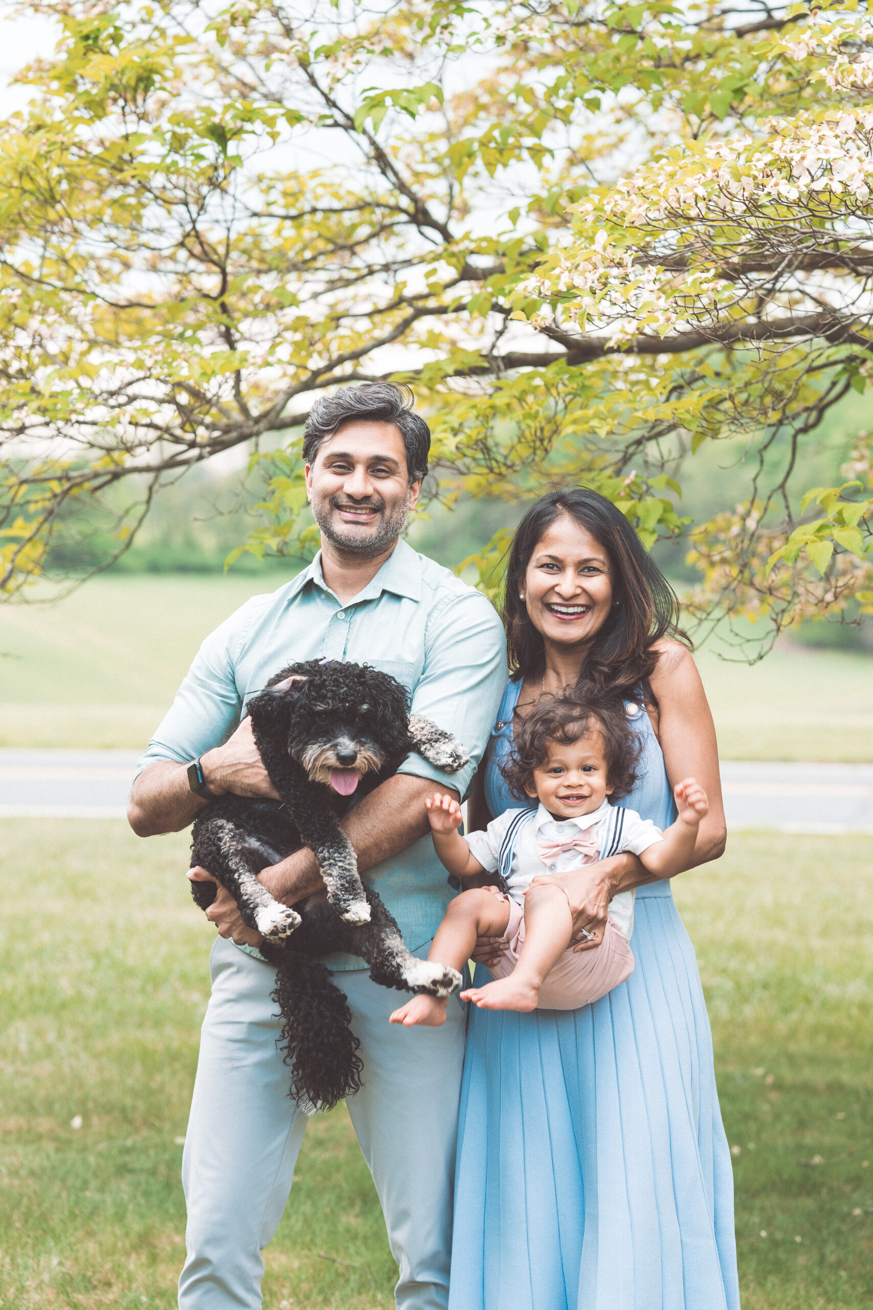 Family of three smiling together while holding their toddler and dog beneath a leafy tree during a Maryland family photoshoot.
