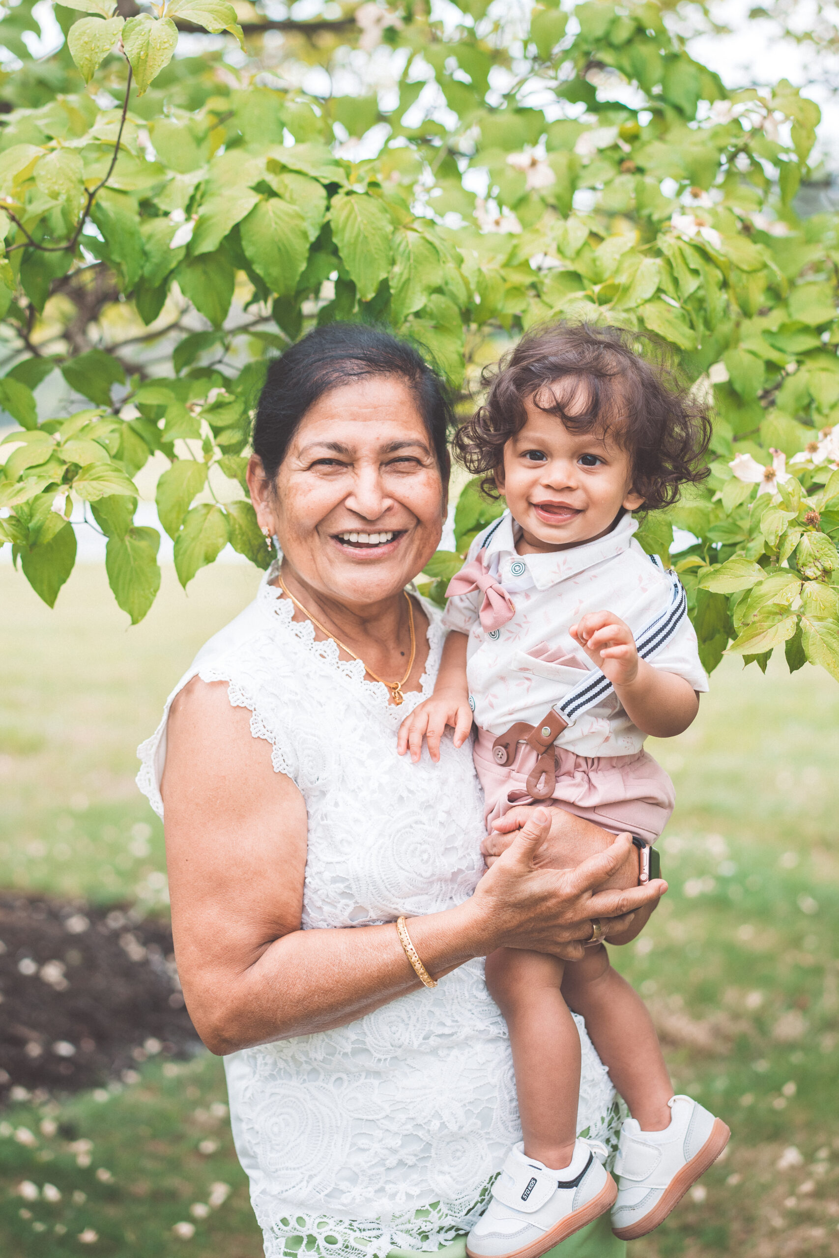 Grandmother holding her smiling great-grandchild beneath green trees during a Maryland family photoshoot.