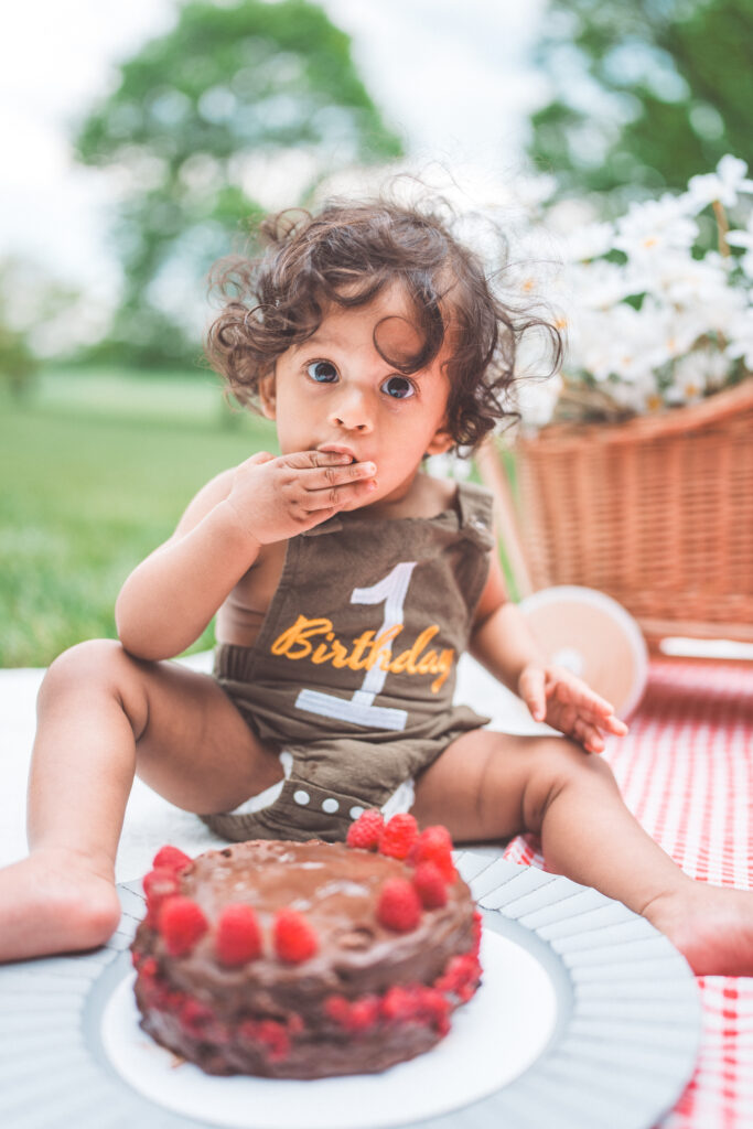 Toddler enjoying a chocolate cake smash while looking up at the camera beside a birthday cake.