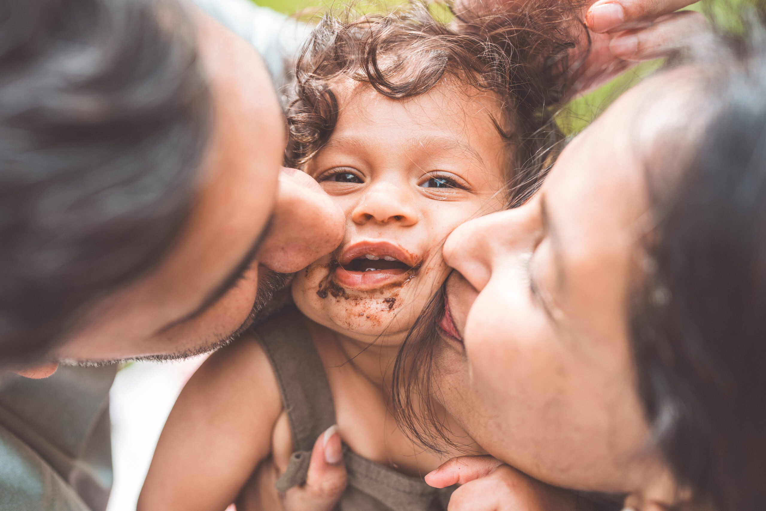 Parents kissing their cake-covered toddler on both cheeks during a playful first birthday celebration.