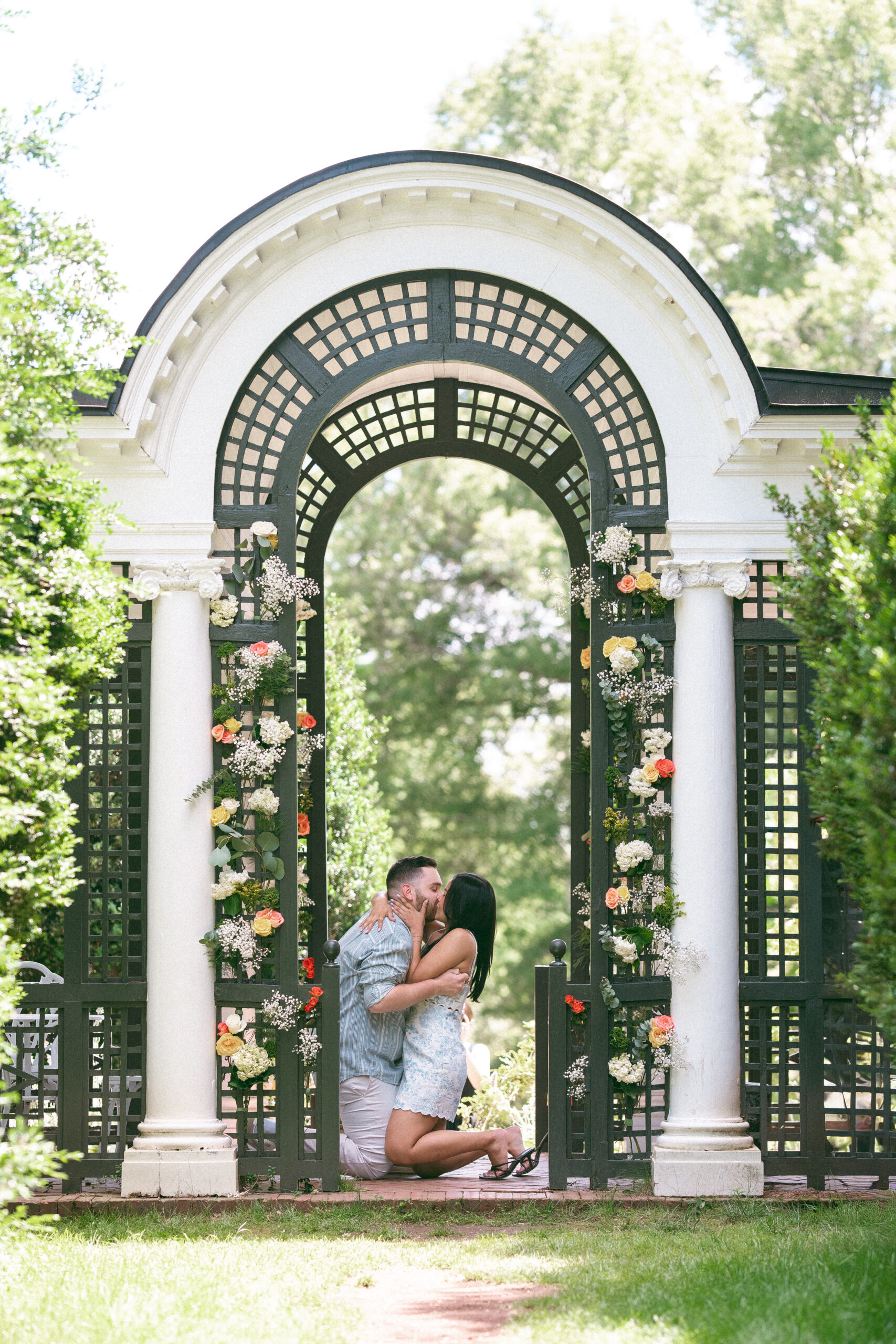 Newly engaged couple sharing a kiss beneath the floral garden arbor.