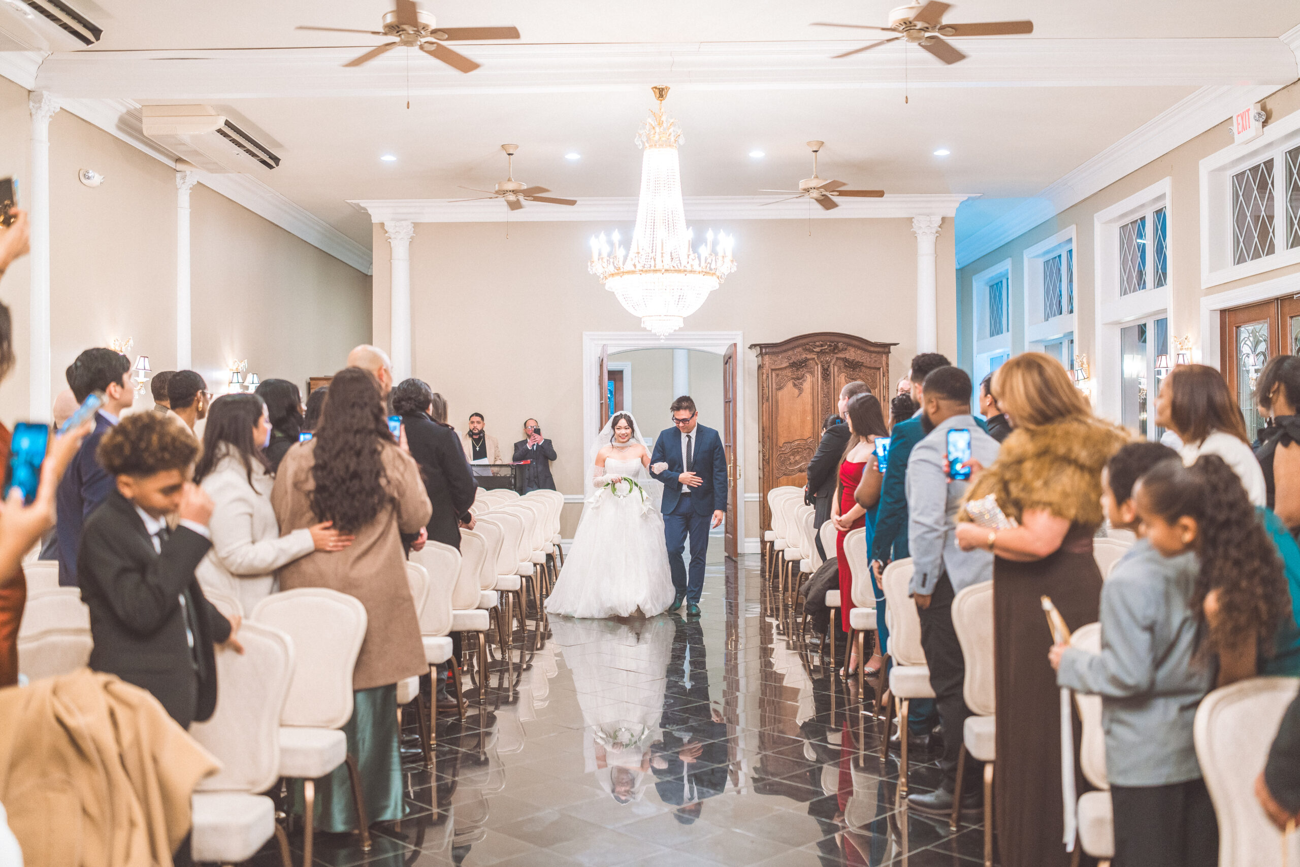 Bride walking down the aisle with her father as wedding guests watch and take photos during the indoor ceremony.