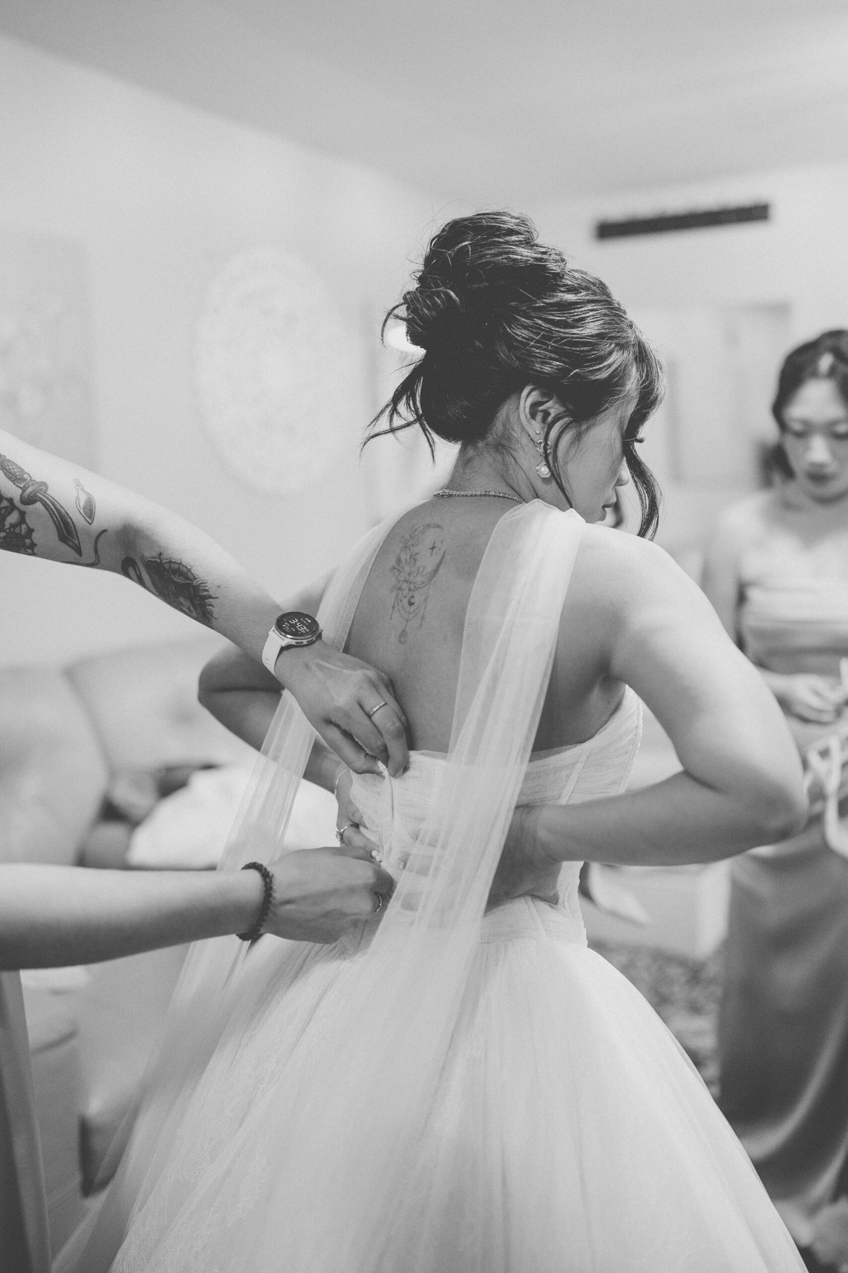 Black-and-white getting-ready photo of the bride having her wedding dress fastened before the ceremony.