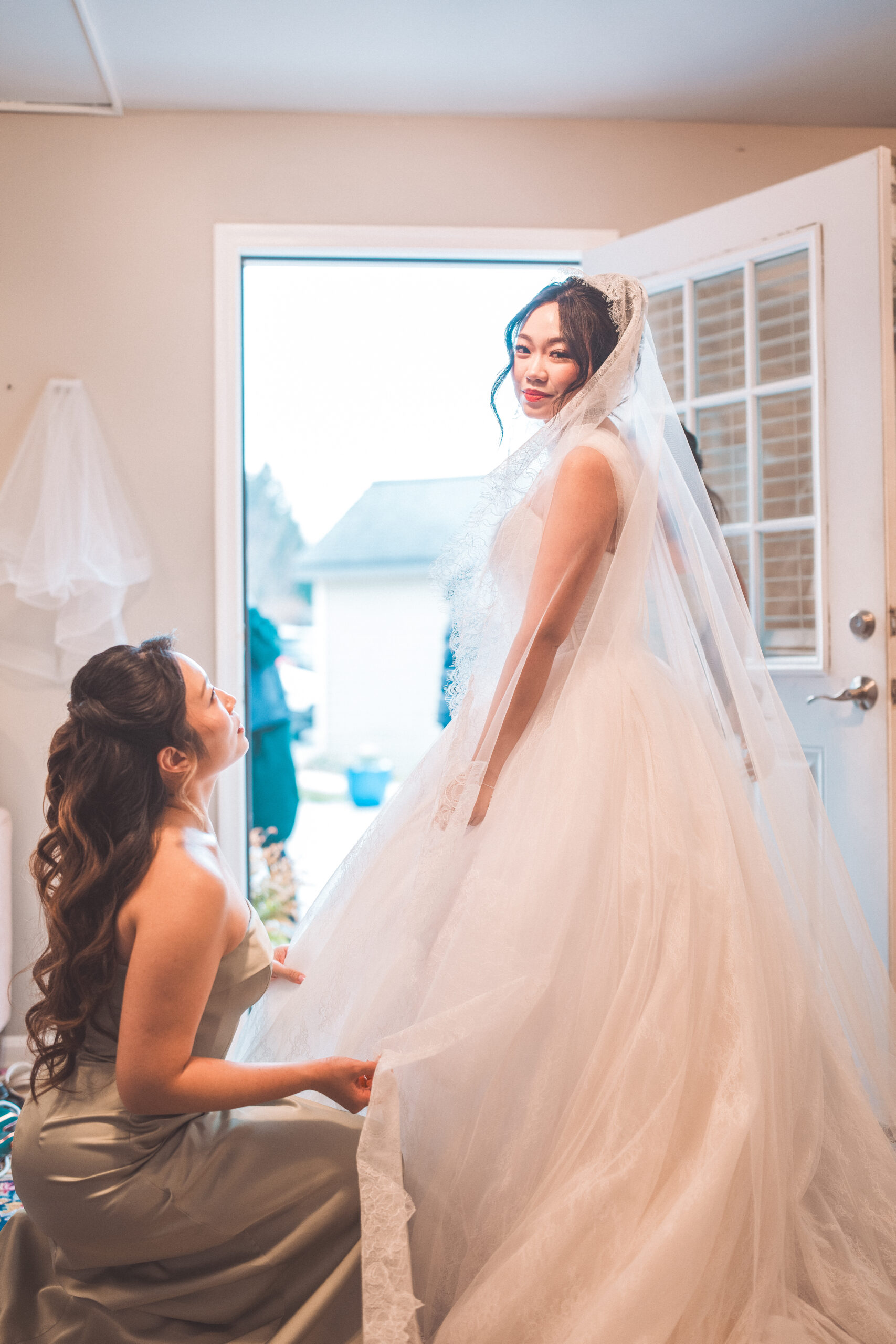 Bride in a lace veil smiling while a bridesmaid adjusts her wedding dress at Cameron Estate Inn.