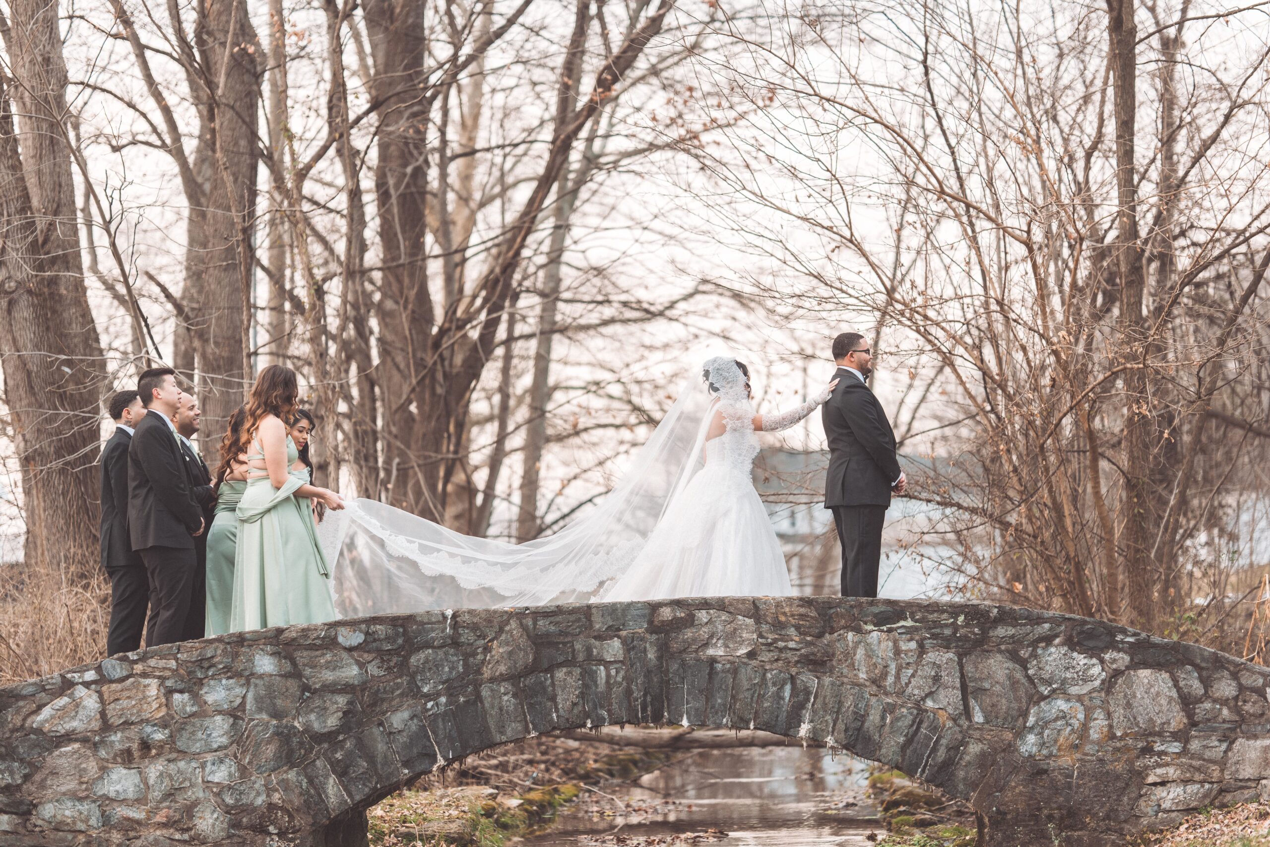 Bride approaching the groom from behind for their first look on a stone bridge while bridesmaids hold her long veil.