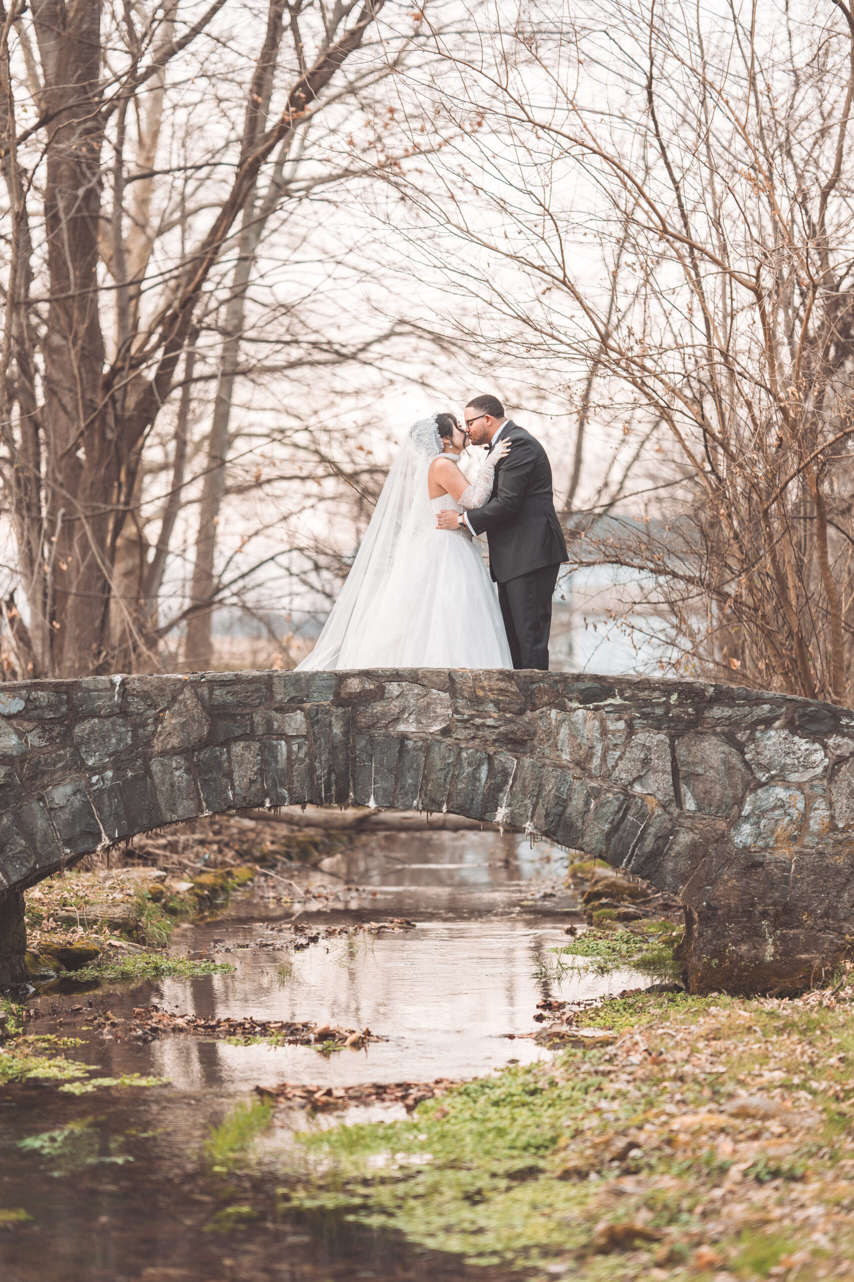 Bride and groom kissing on a stone bridge surrounded by bare trees and a quiet creek.